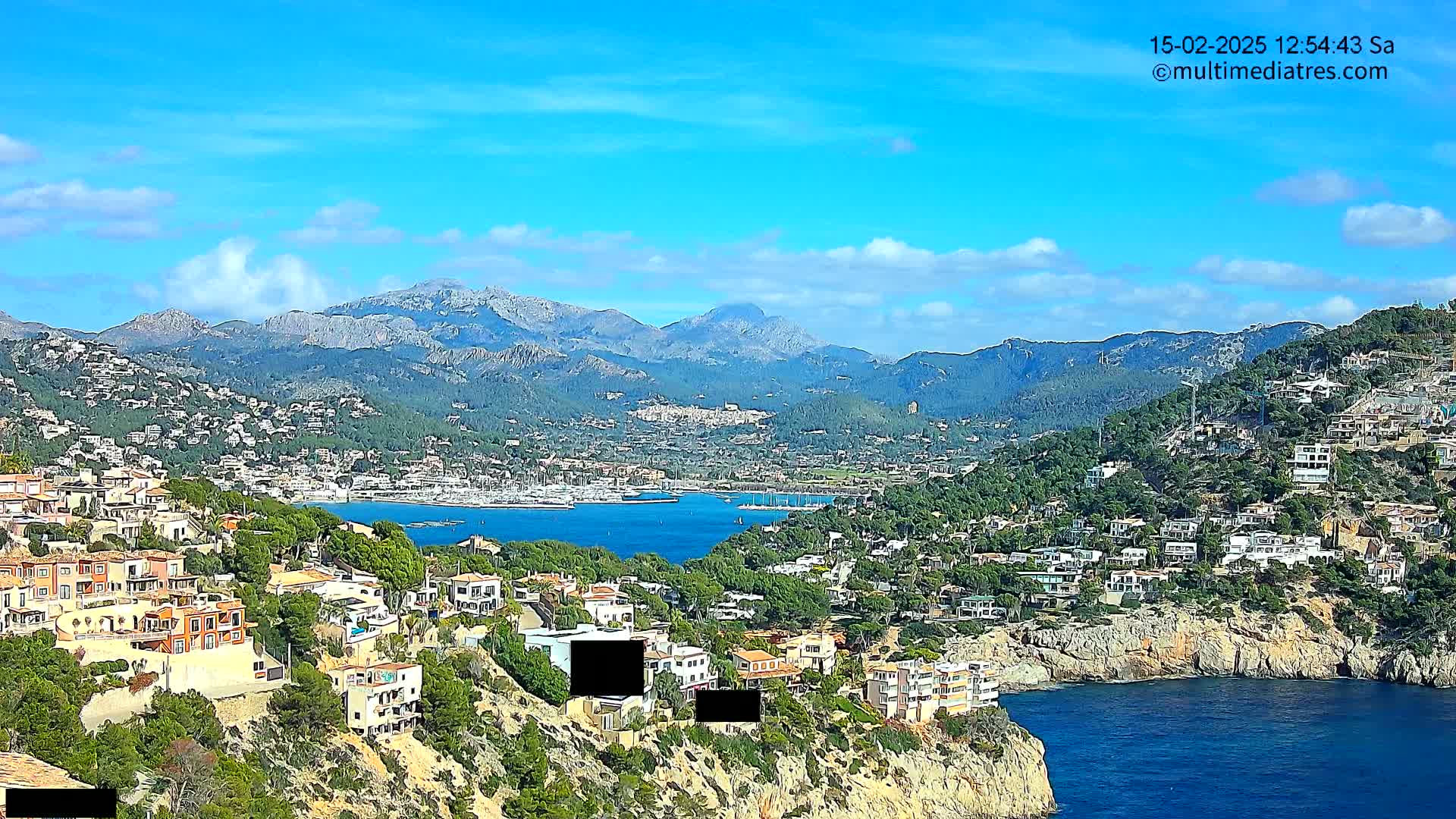 A coastal town nestled among green hills and mountains under a mostly clear, bright blue sky overlooks a calm blue bay filled with boats.