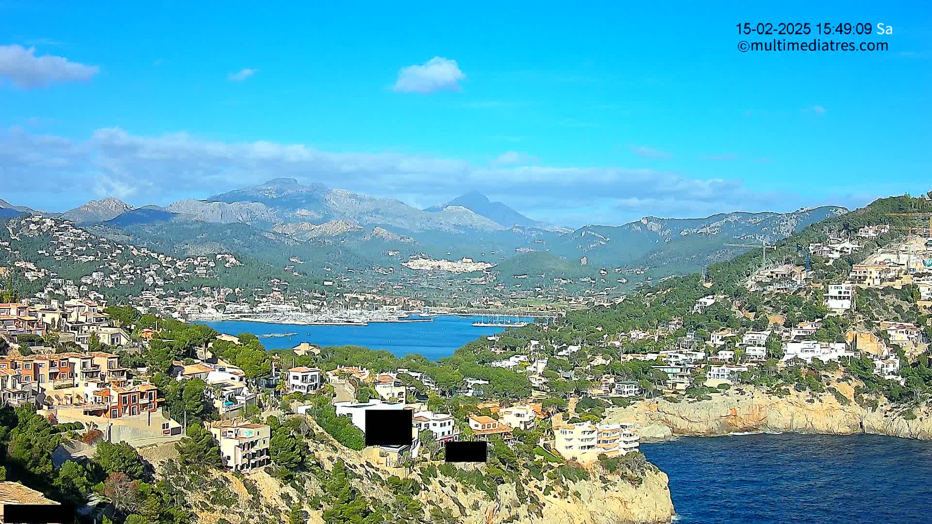 A coastal town nestled on a hillside overlooking a calm bay, with mountains in the background under a mostly clear blue sky.