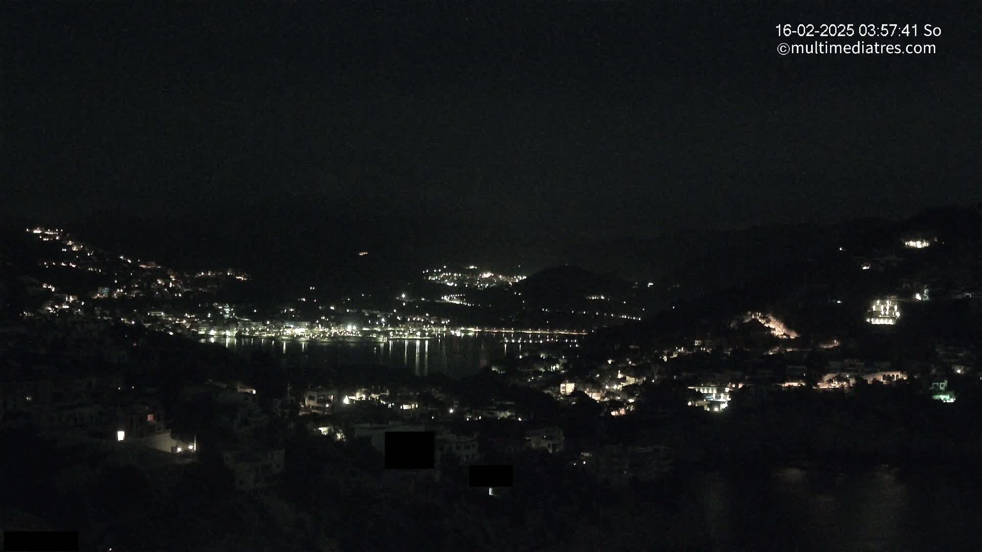A nighttime view of a coastal town nestled on hillsides, with its lights reflecting on a calm body of water under a clear, dark sky.