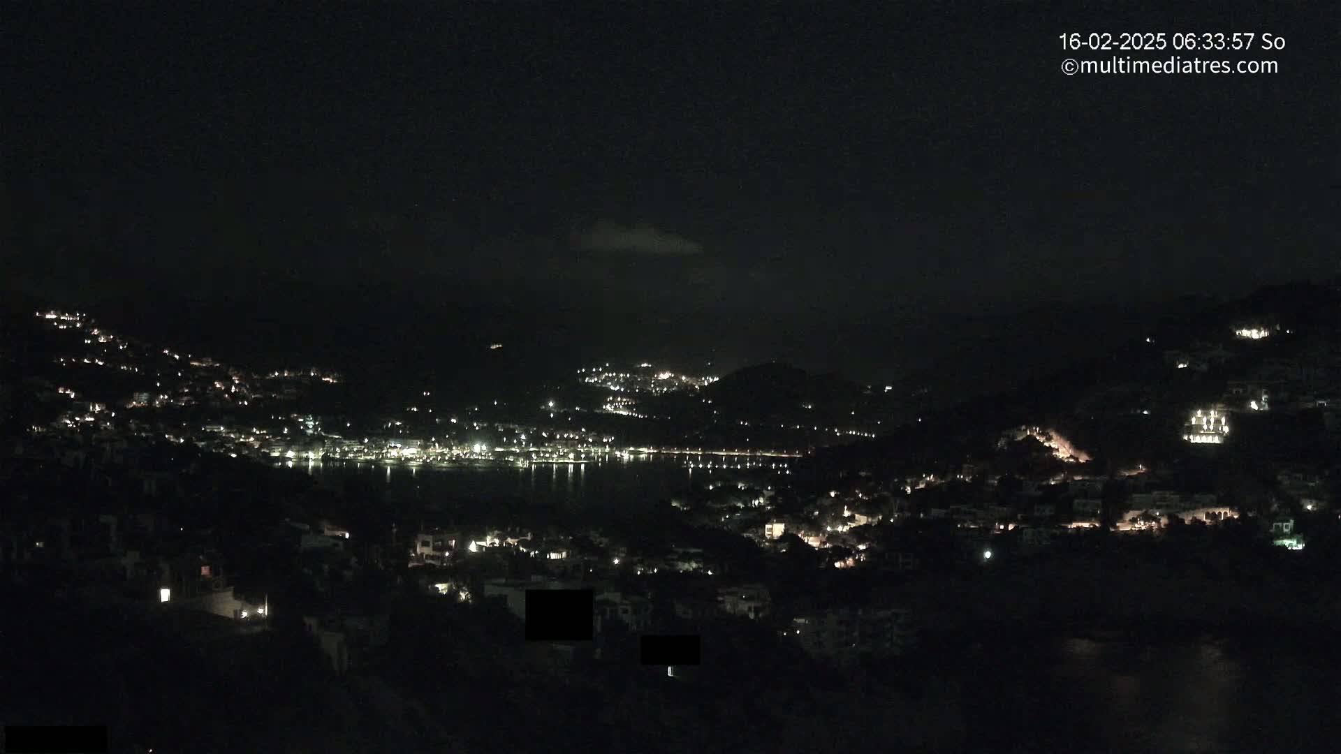 A nighttime view of a coastal town nestled on hillsides, with scattered lights illuminating the buildings and a dark body of water in between.