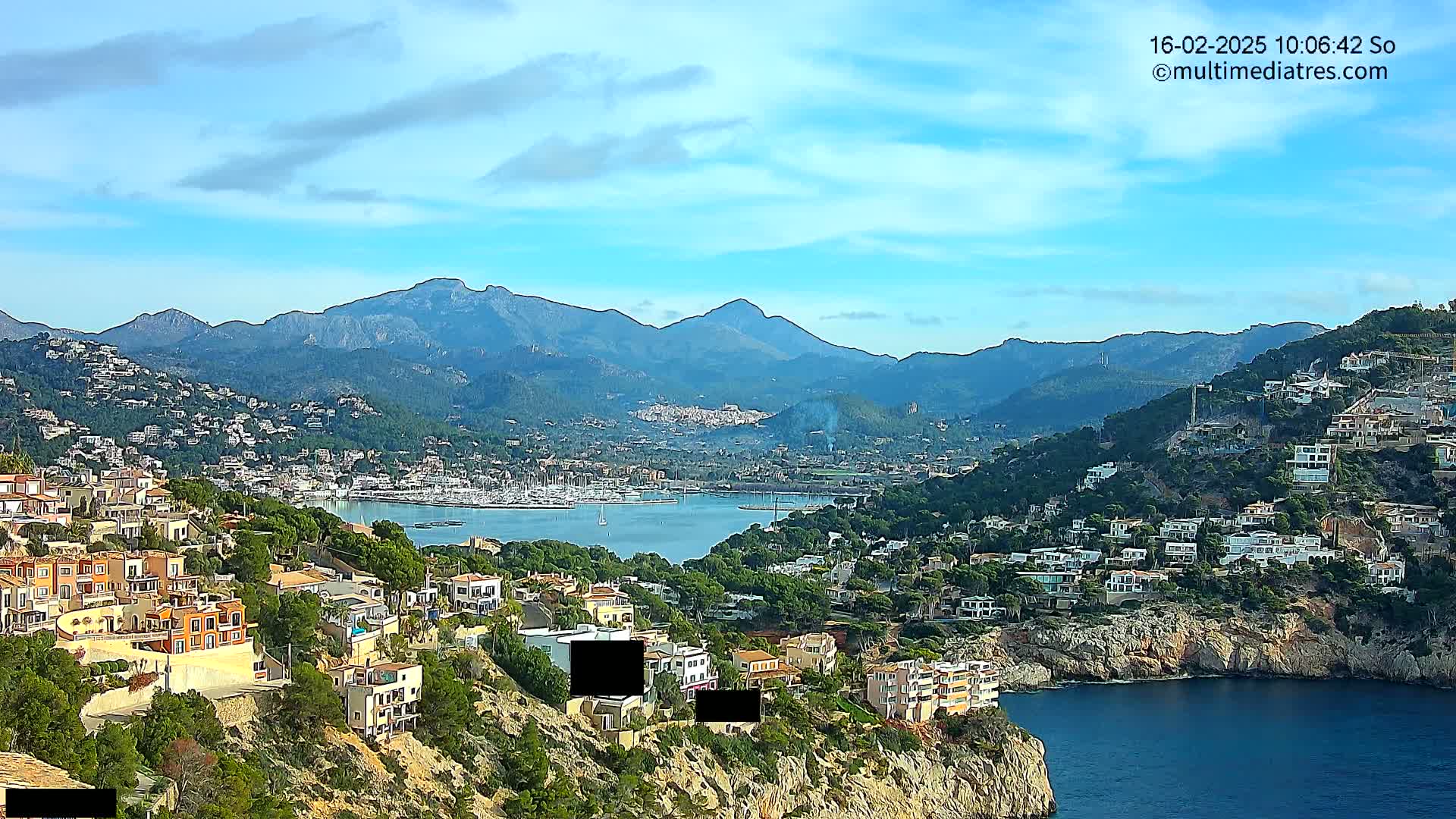 A coastal town nestled against a mountain range under a partly cloudy sky, featuring a harbor filled with boats and numerous houses built into the hillsides.