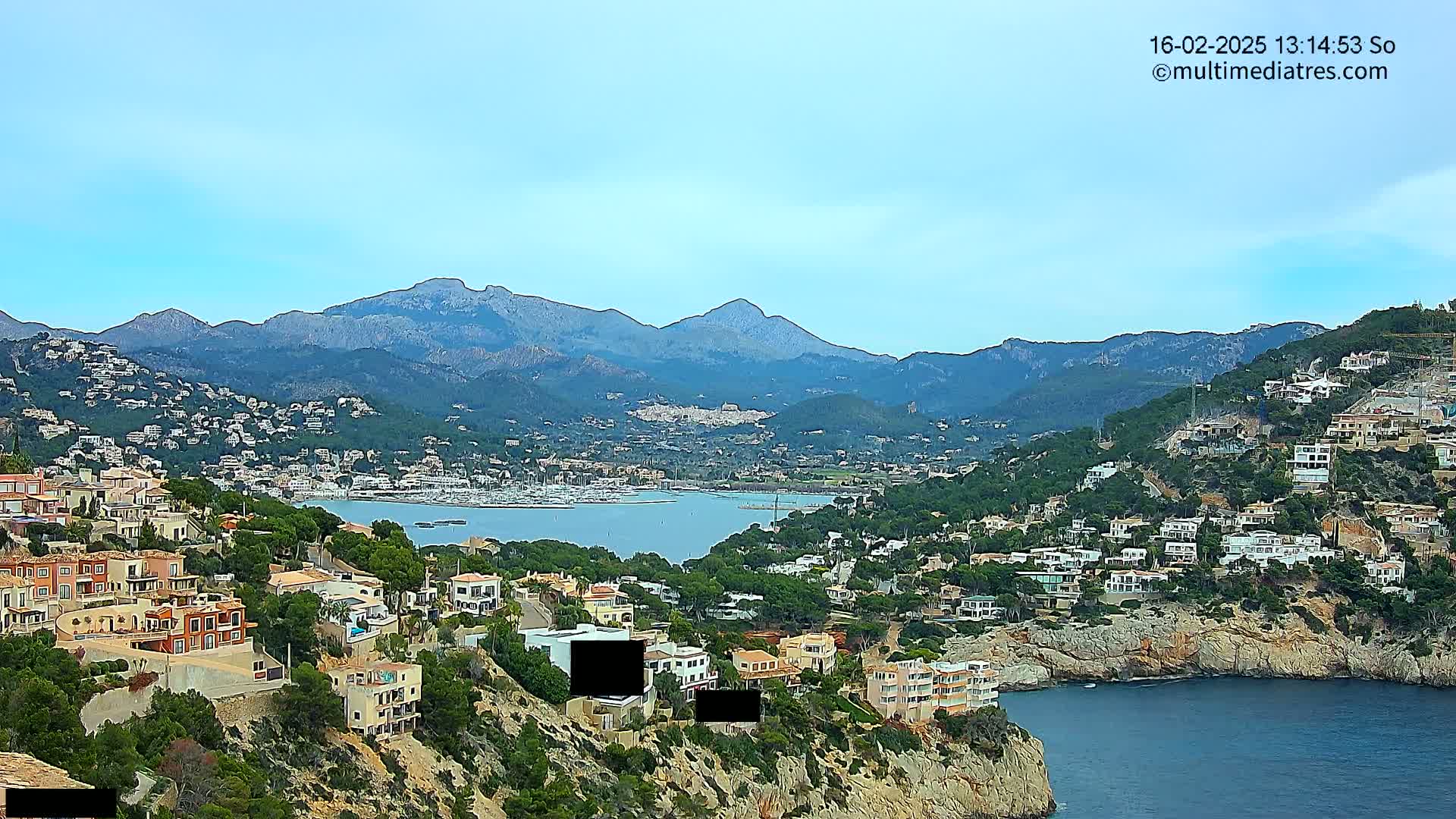 A coastal town nestled in a hillside, overlooking a calm bay and a mountain range under a clear sky.