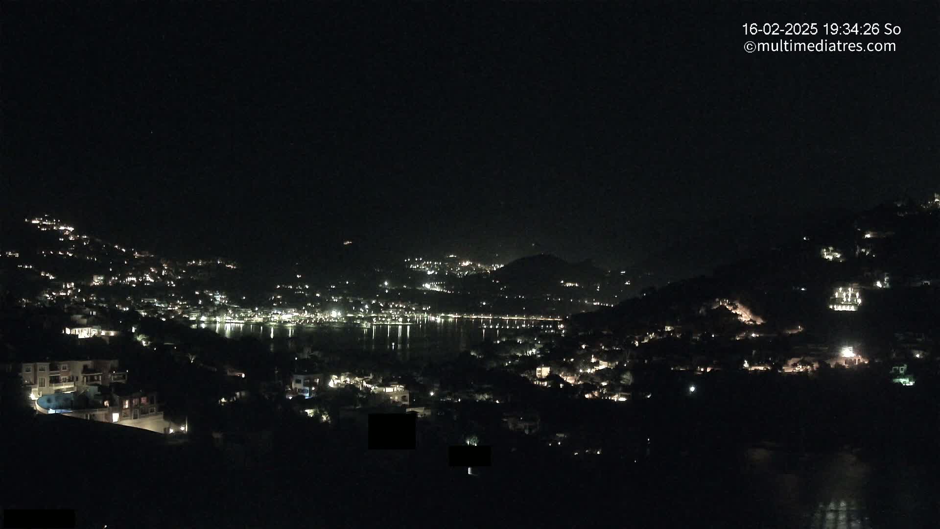 A nighttime view of a coastal town nestled on hillsides, with scattered lights illuminating the buildings and reflecting on the calm water below under a clear, dark sky.