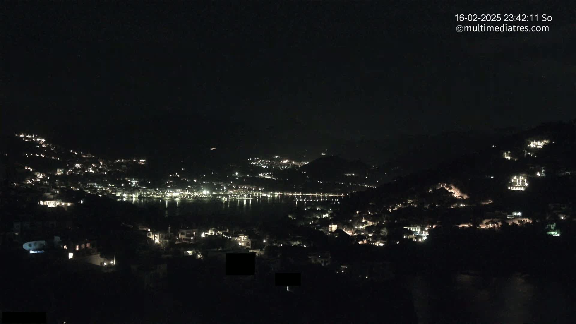 A nighttime view shows a coastal town nestled on hillsides, its lights twinkling against a dark sky over a calm body of water.