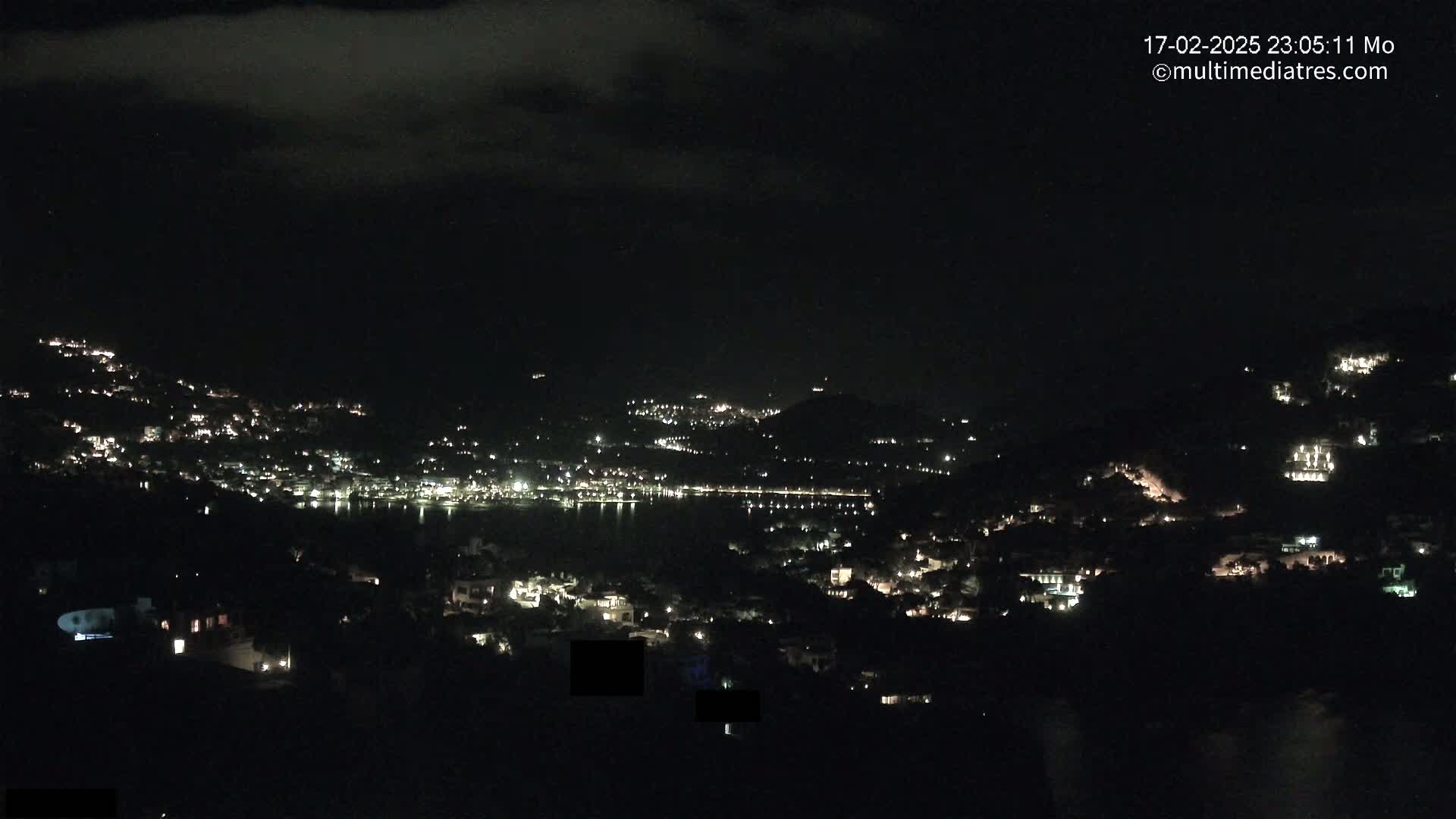 A nighttime view of a coastal town illuminated by scattered lights, nestled on hillsides overlooking a dark body of water under a cloudy sky.
