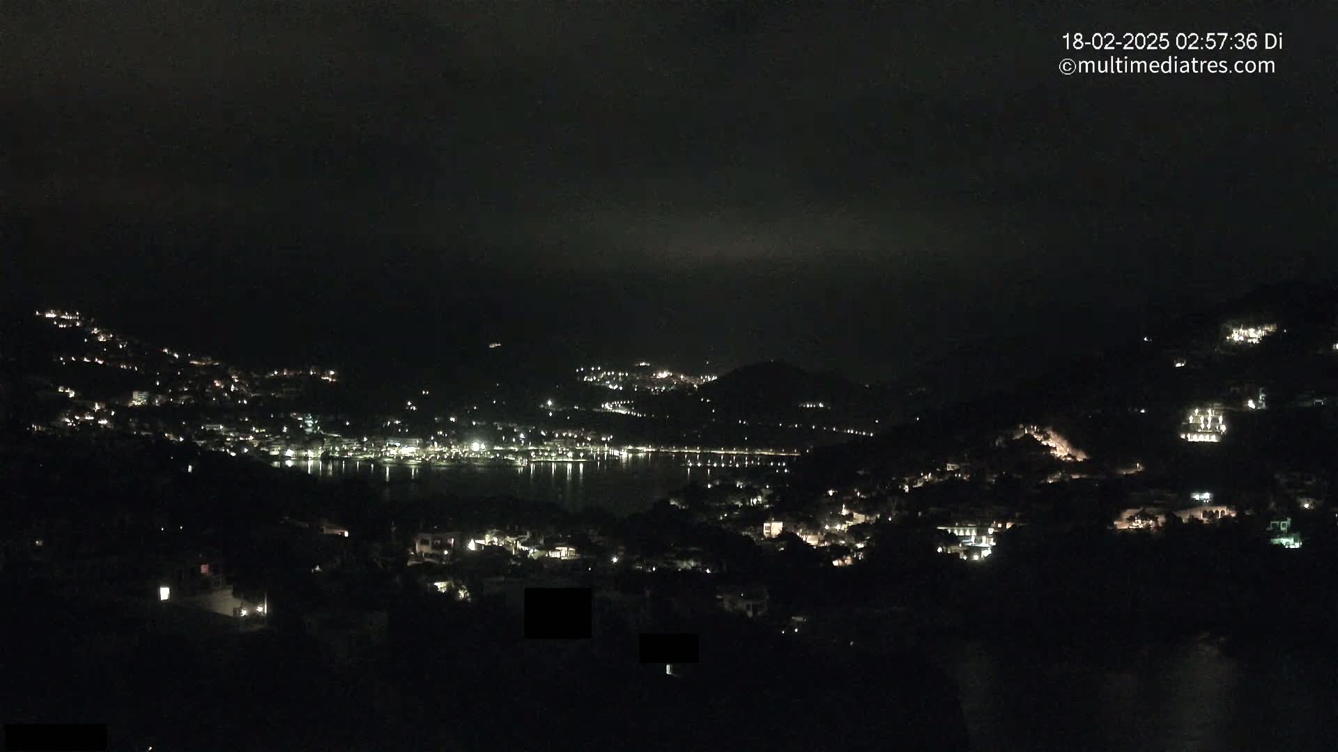 A nighttime view of a coastal town nestled in hills, with scattered lights illuminating the buildings and a dark, calm body of water reflecting the lights.
