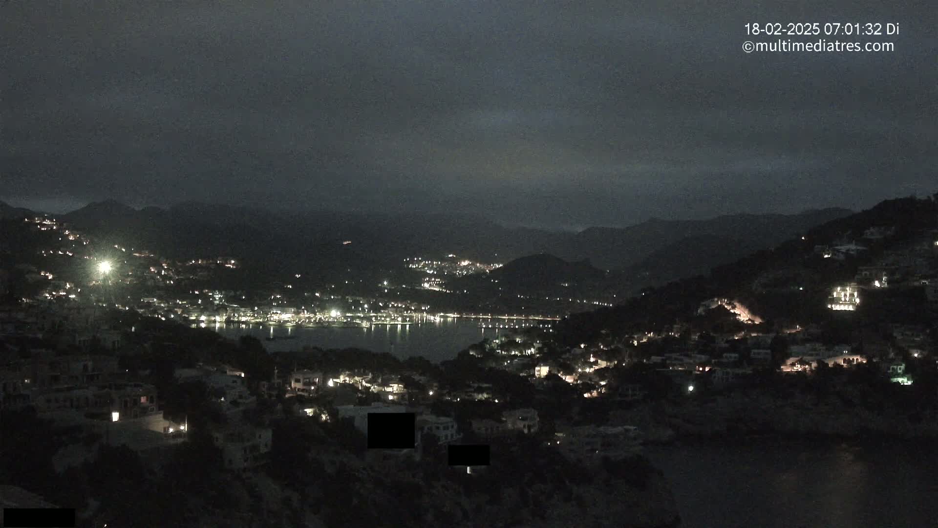 A nighttime view of a coastal town nestled in dark hills, illuminated by scattered lights, under a cloudy sky.