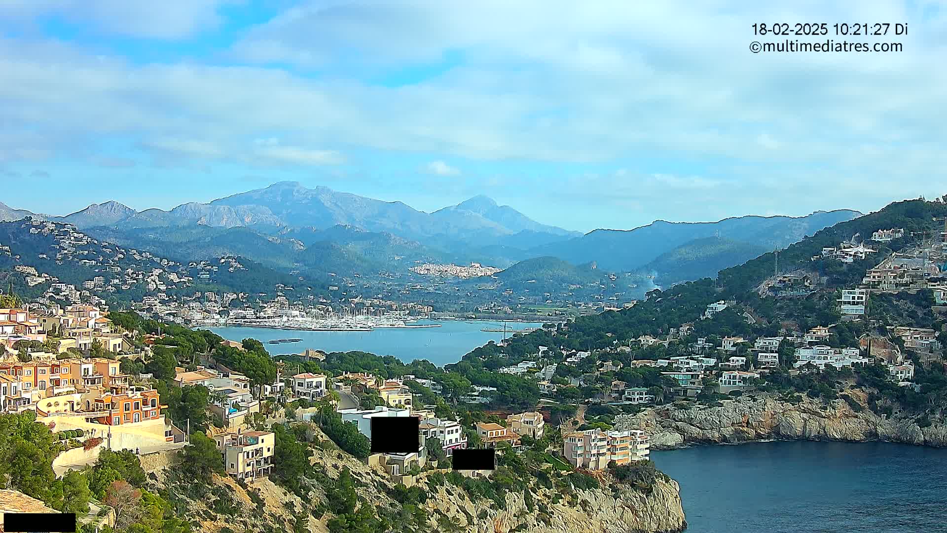 A coastal town nestled in a mountainous region overlooks a calm bay filled with boats under a partly cloudy sky.