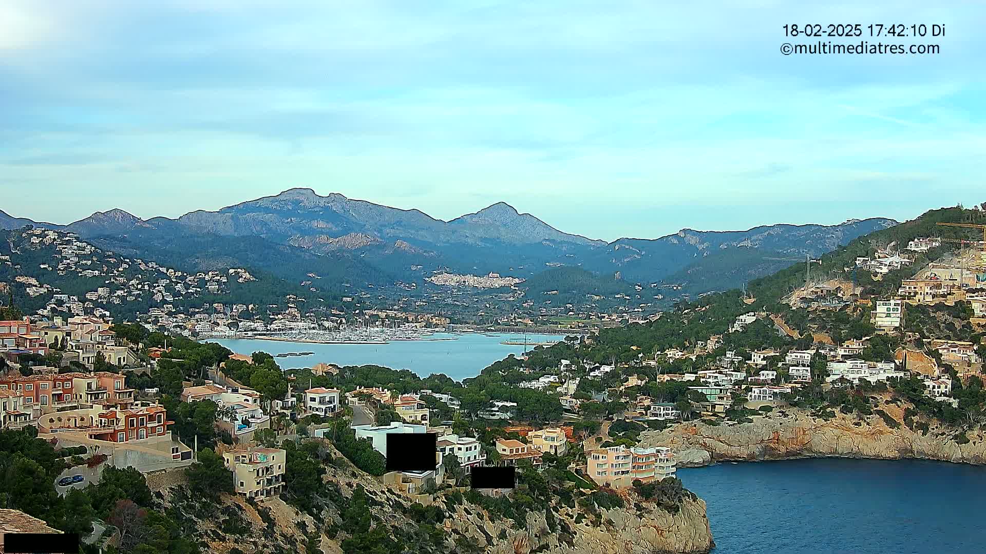 A coastal town nestled in a mountainous region under a mostly clear sky, featuring a calm bay with boats and numerous houses built into the hillsides.