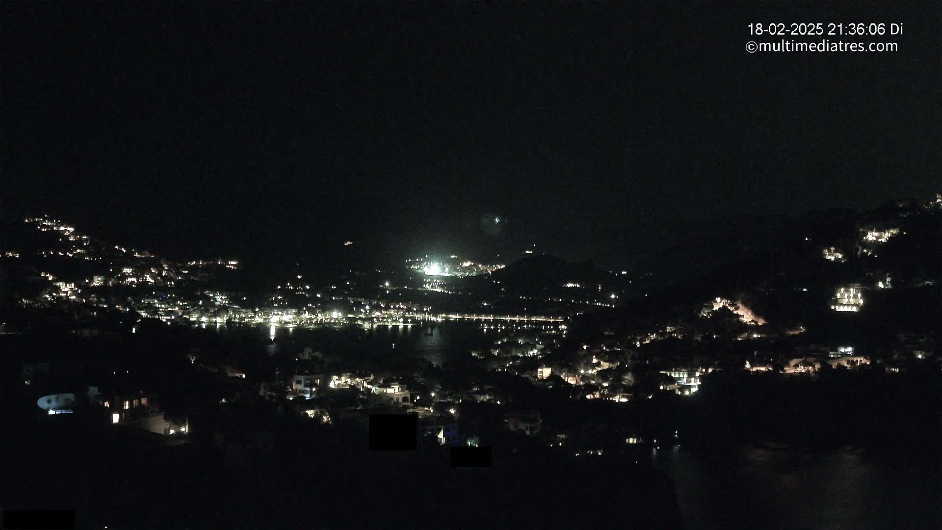 A nighttime view of a coastal town illuminated by scattered lights, situated on hillsides overlooking a dark body of water under a clear, dark sky.