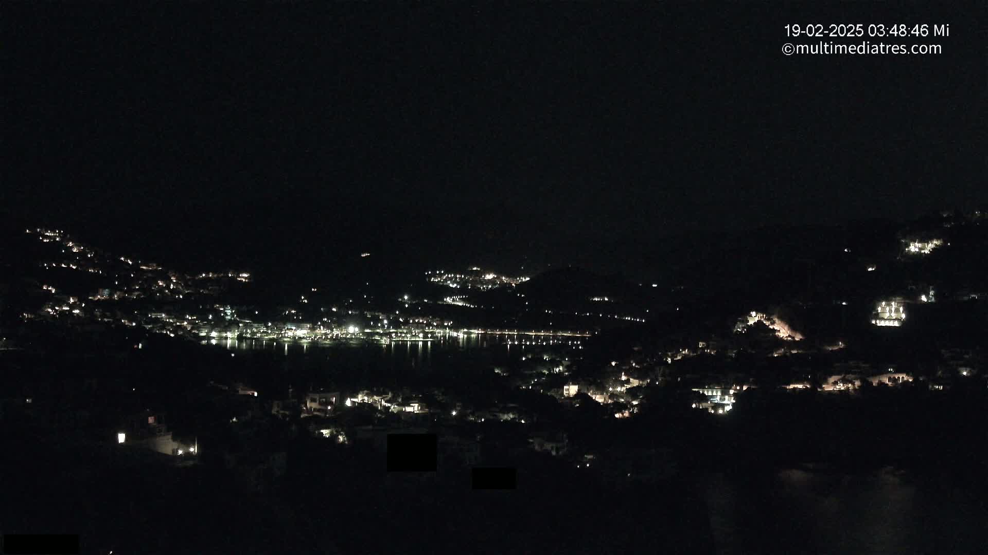 A nighttime view of a coastal town illuminated by scattered lights, with a dark body of water visible in the middle distance.
