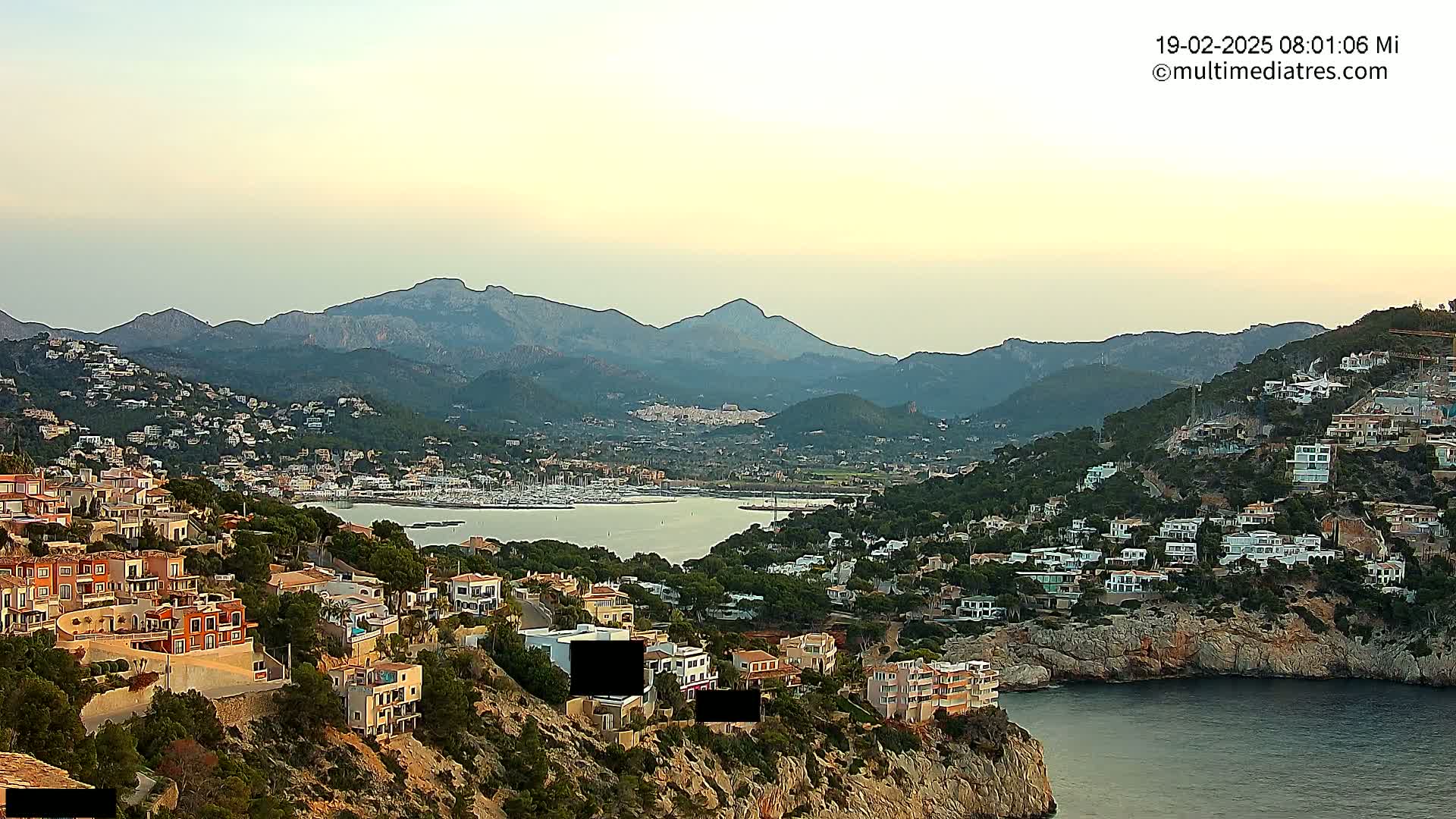 A coastal town nestled into a hillside overlooks a calm bay and distant mountains under a pale sunset sky.