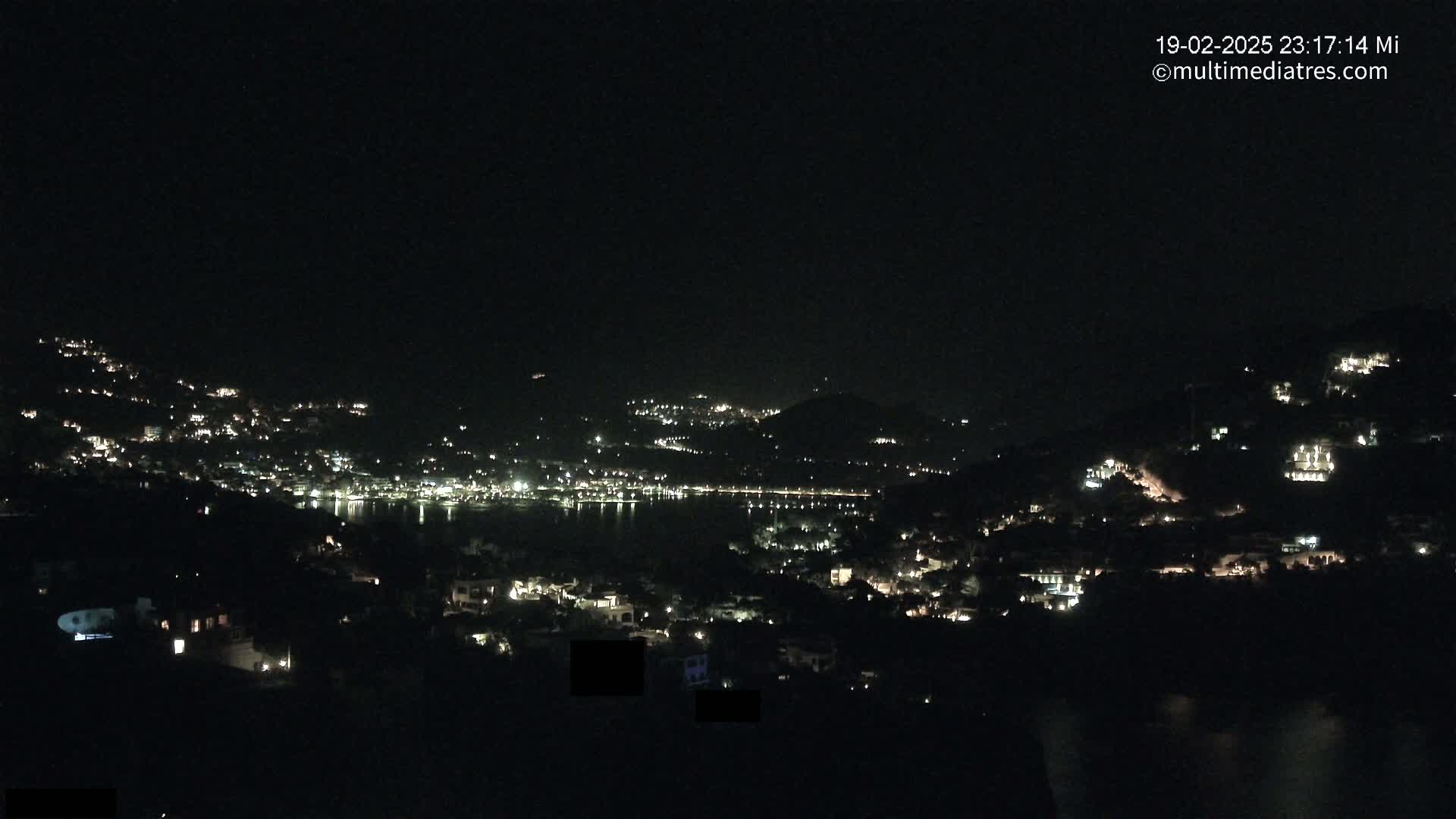A nighttime view of a coastal town nestled on hillsides, illuminated by scattered lights, with a dark body of water visible in the distance.