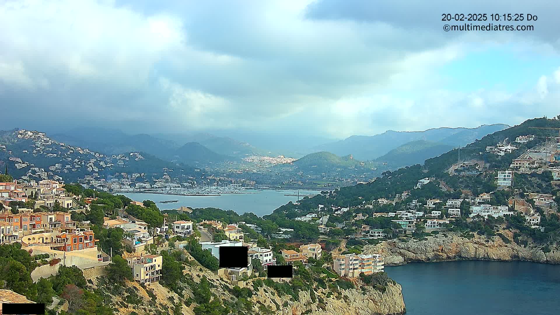 A coastal town nestled on a hillside overlooking a calm bay dotted with boats, under a partly cloudy sky.