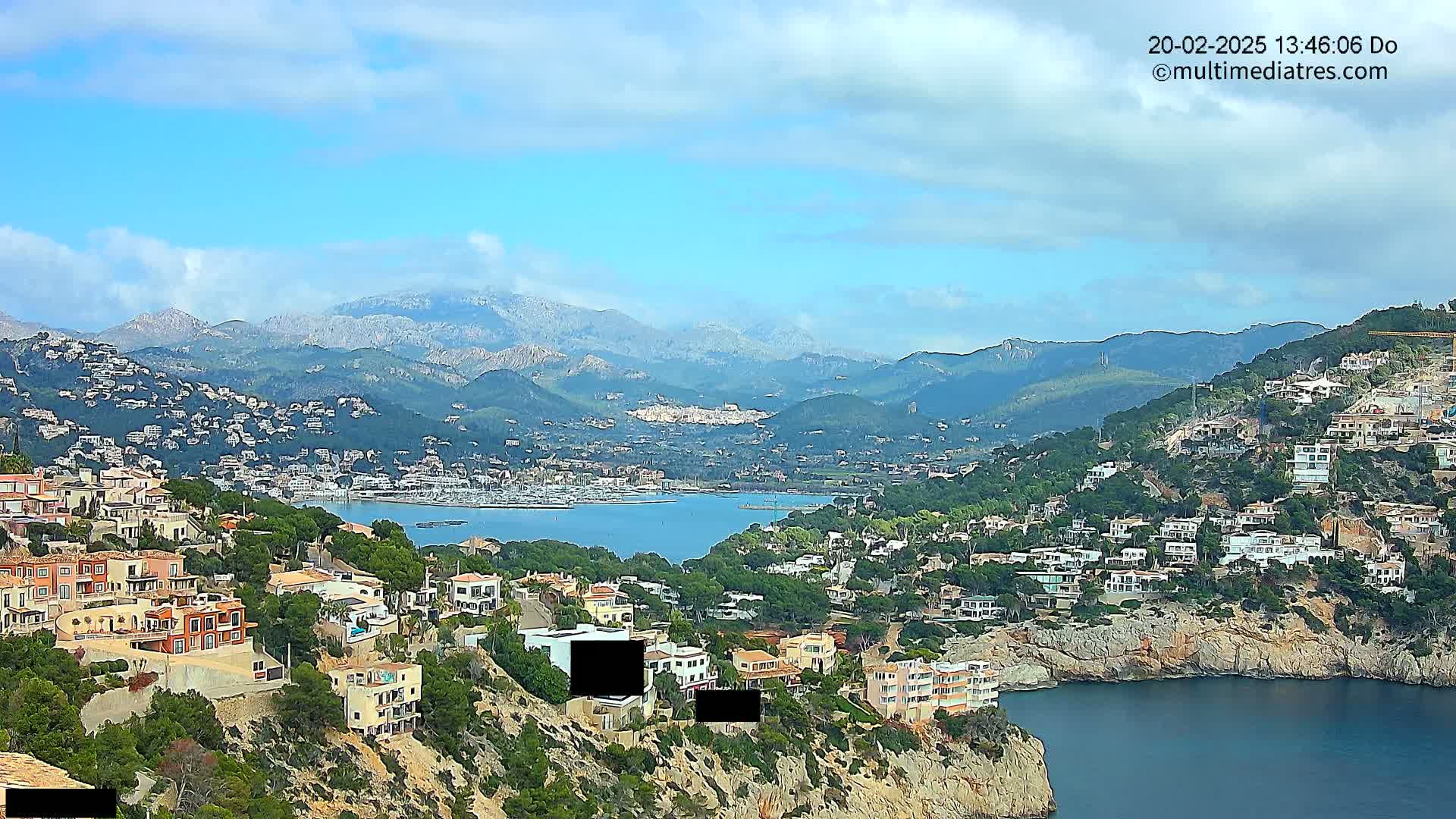 A coastal town nestled in hills overlooking a calm bay, with snow-capped mountains in the distance under a partly cloudy sky.
