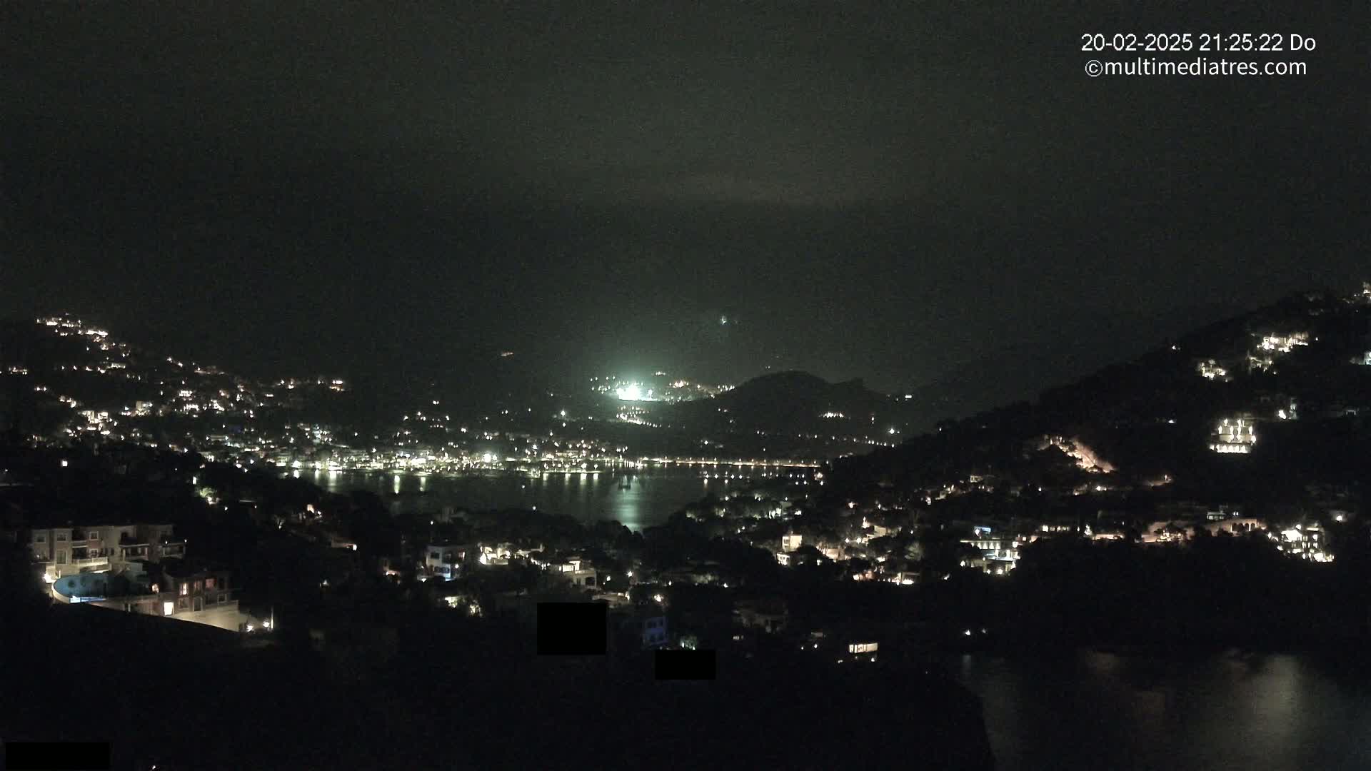 A nighttime view of a coastal town nestled on hillsides surrounding a dark body of water, under a mostly clear sky.