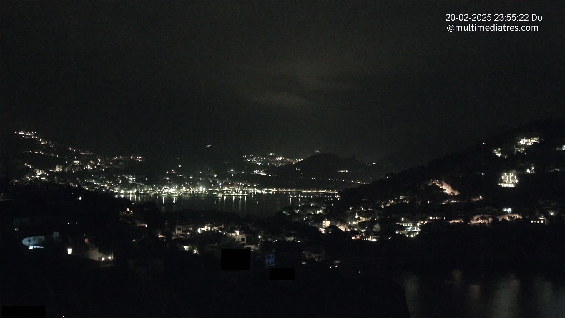 A nighttime view of a coastal town nestled on hillsides, illuminated by scattered lights, with a calm body of water reflecting the lights.