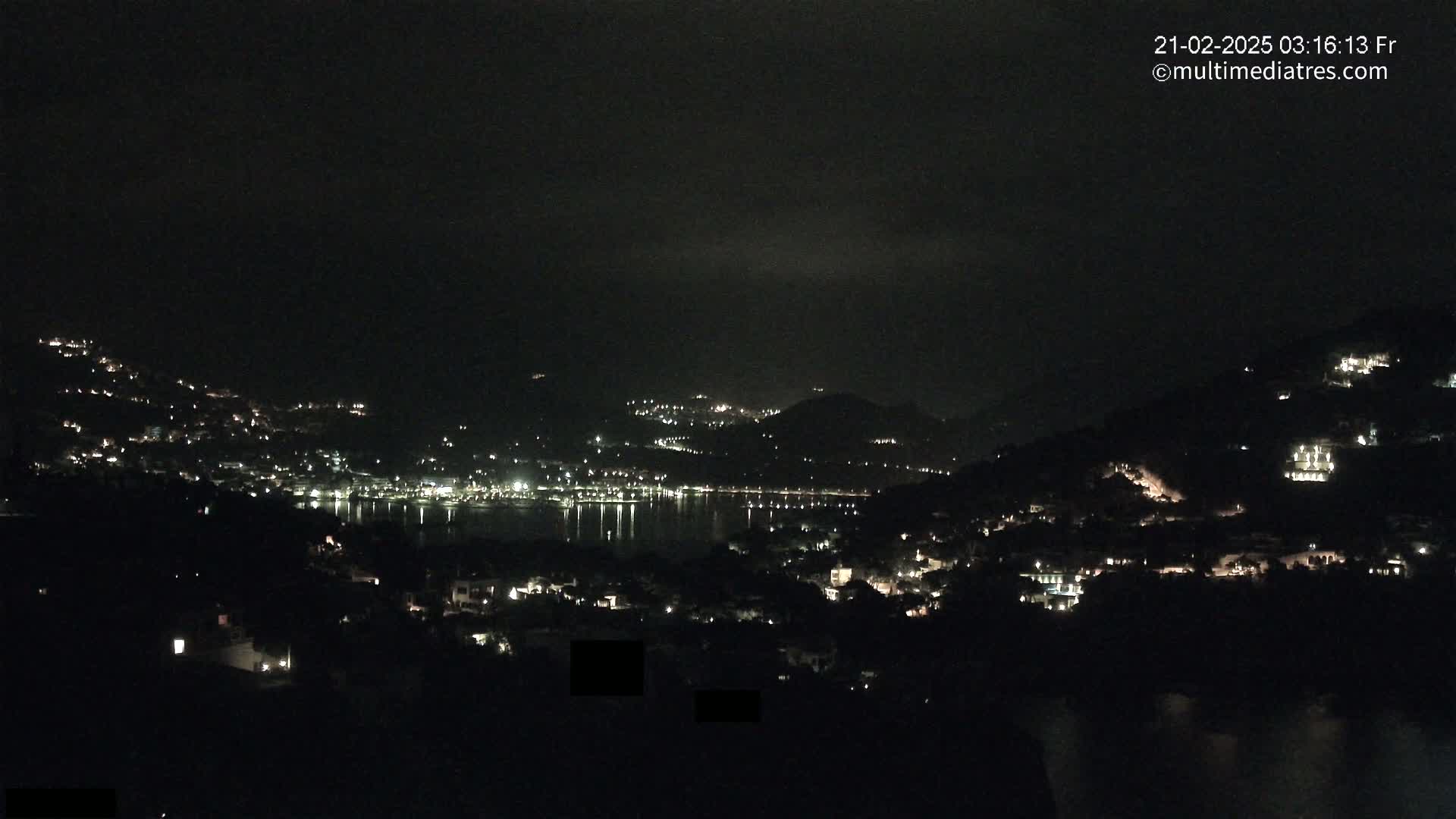 A nighttime view of a coastal town nestled on hillsides, with scattered lights reflecting on the water below under a dark, possibly cloudy sky.