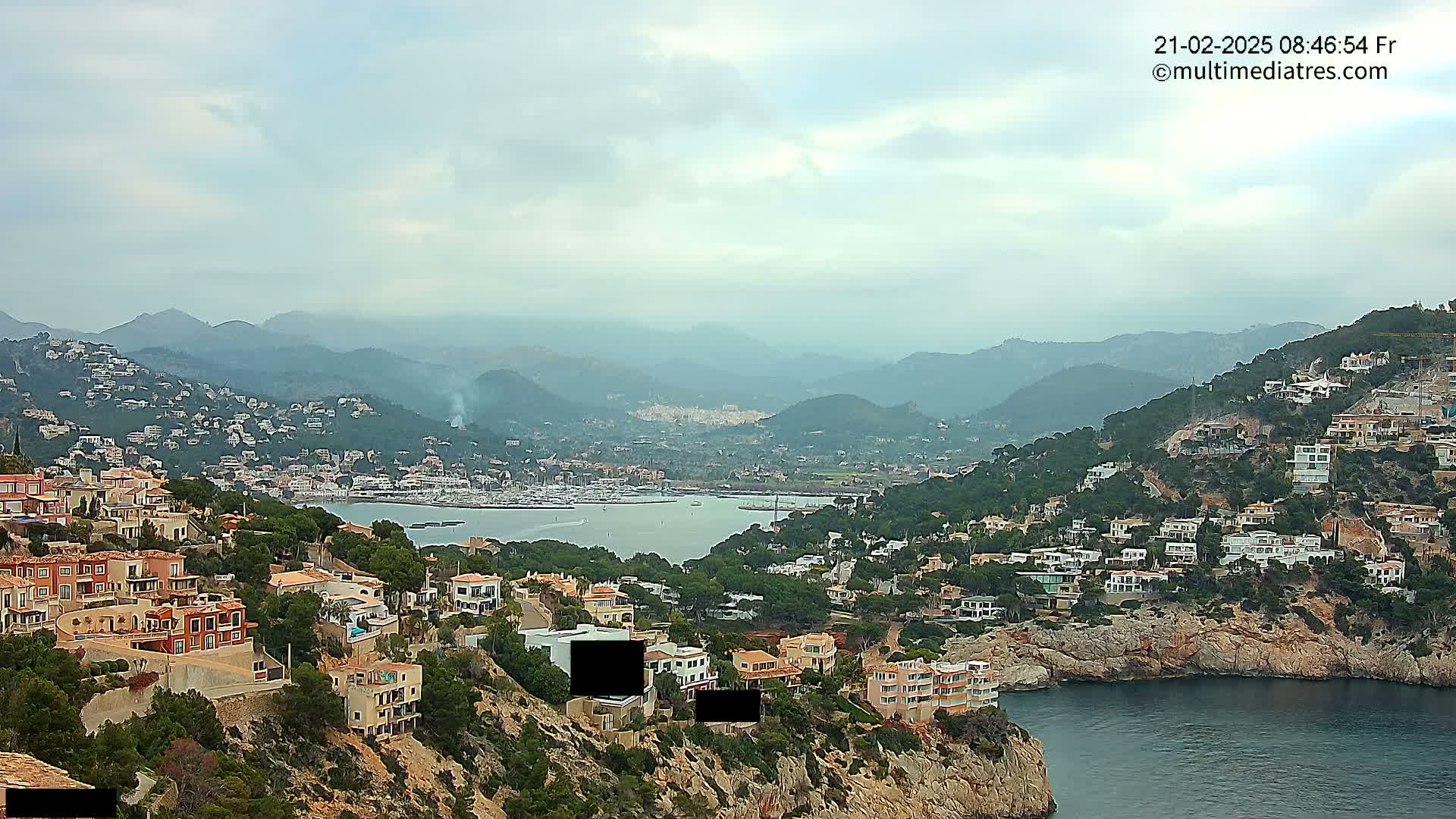 A coastal town nestled in a mountainous region under a cloudy sky, with a calm bay filled with boats in the foreground.