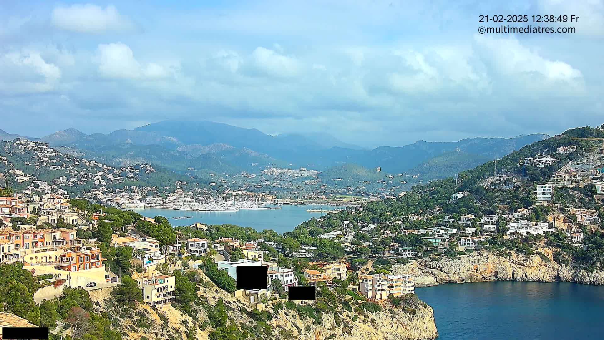 A coastal town nestled among mountains under a partly cloudy sky, with a calm bay filled with boats in the foreground.