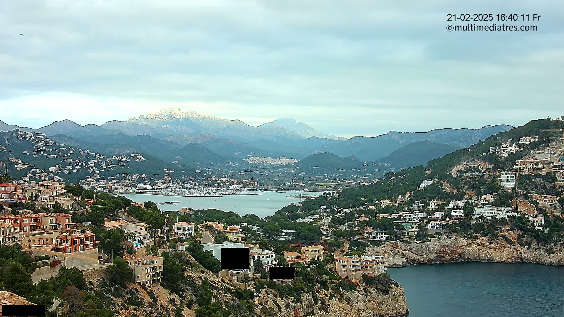 A coastal town nestled in a mountainous region, with a calm bay and snow-capped mountains in the background under a cloudy sky.