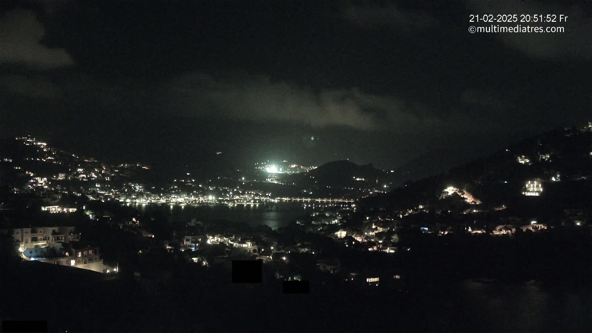 A nighttime view of a coastal town nestled on hillsides, with scattered lights illuminating the buildings and a dark body of water in the center.