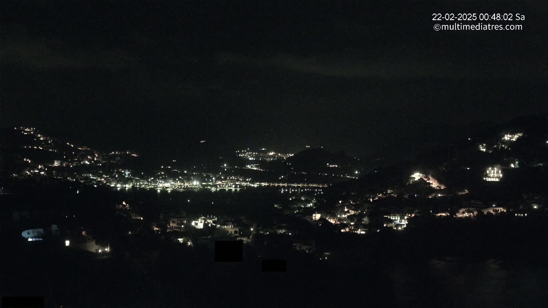 A nighttime view of a hillside town illuminated by scattered lights under a dark, cloudy sky.