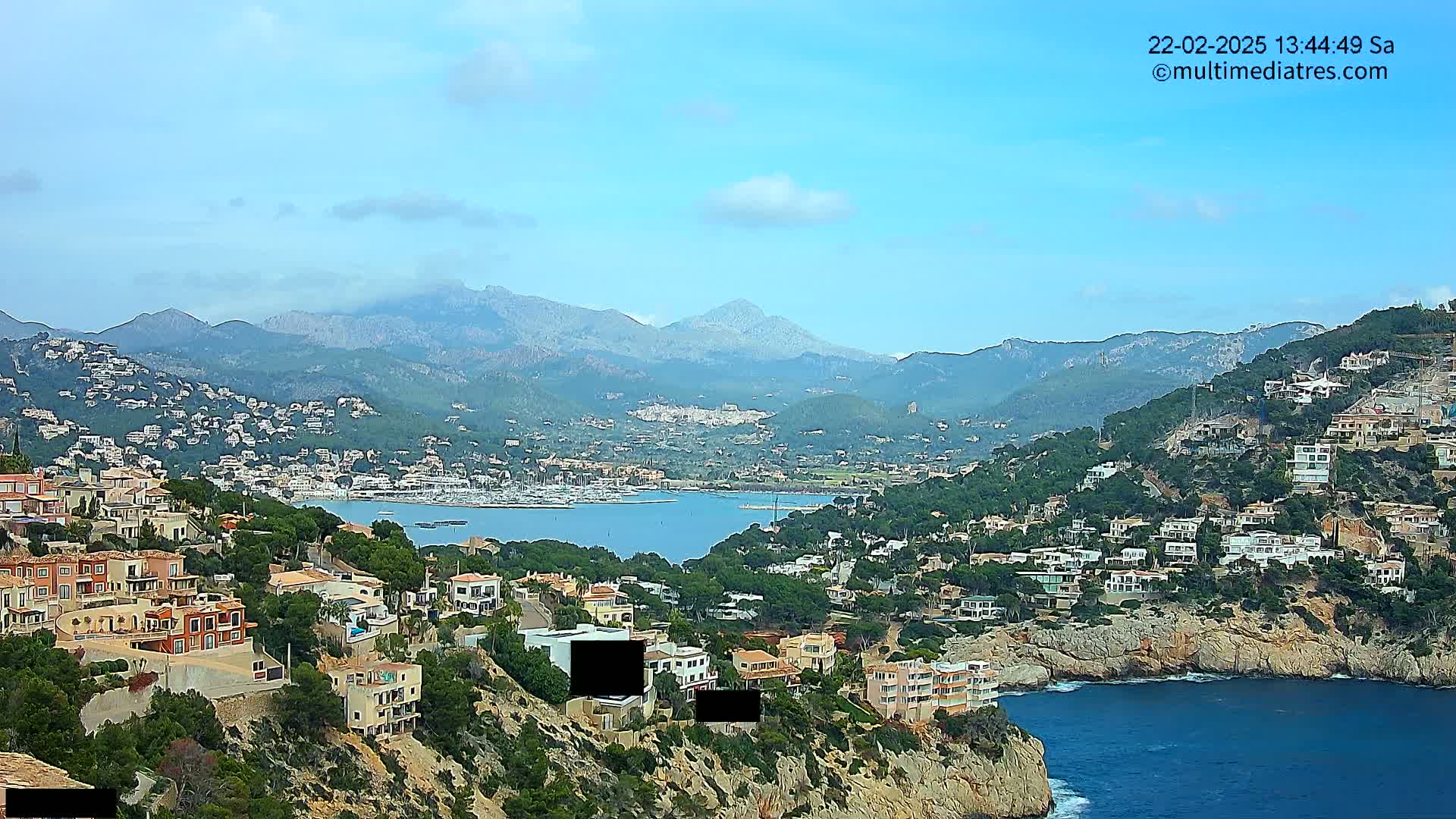 A coastal town nestled on a hillside overlooking a calm bay and mountains under a mostly clear sky.