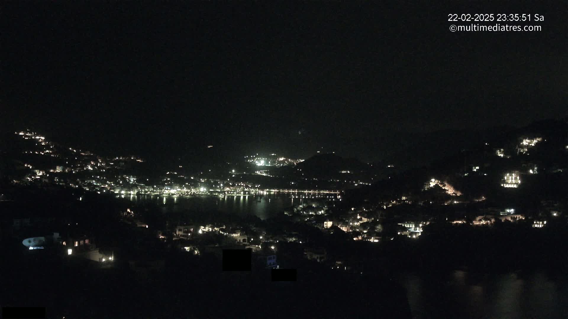 A nighttime view of a coastal town nestled on hillsides overlooking a dark body of water, with scattered lights illuminating buildings and streets.