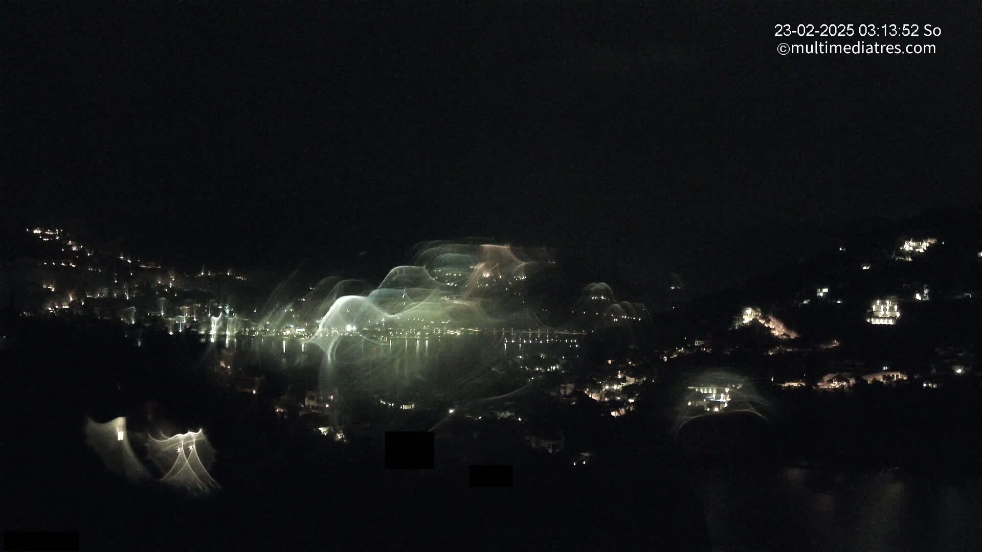 A nighttime view of a town nestled on hillsides overlooking a body of water, with lights illuminating the buildings and faint, streaky lights possibly from water features or long exposure photography reflecting on the water.