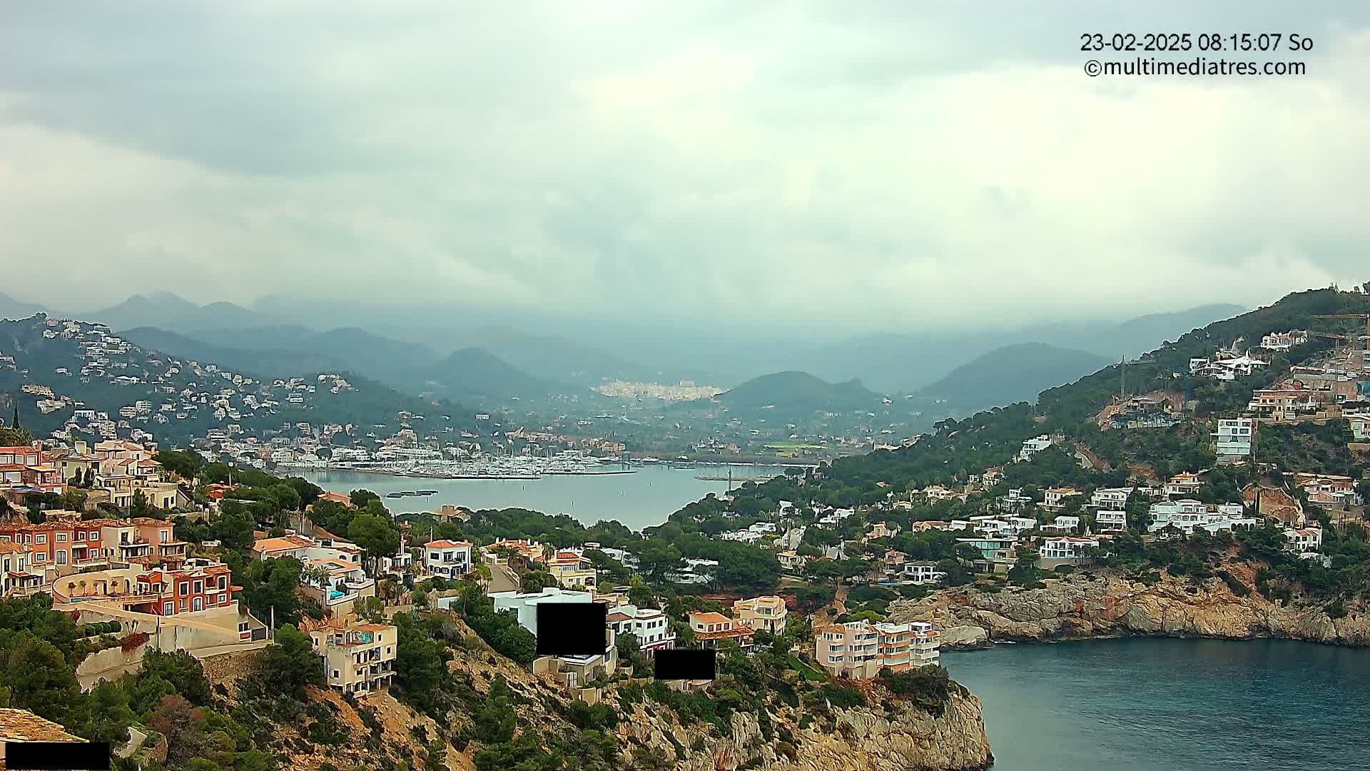 A coastal town nestled in hills overlooking a calm bay filled with boats under an overcast sky.
