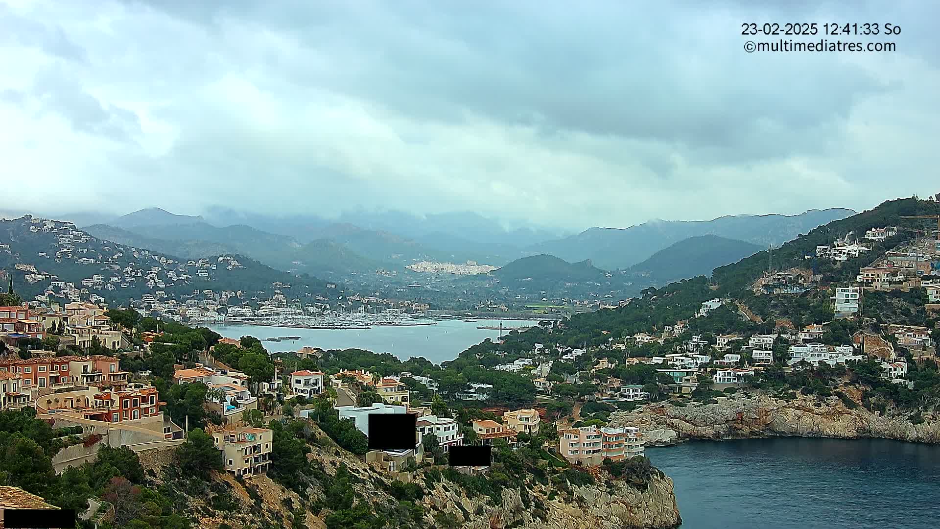 A coastal town nestled in hills overlooking a calm bay filled with boats under a cloudy sky.