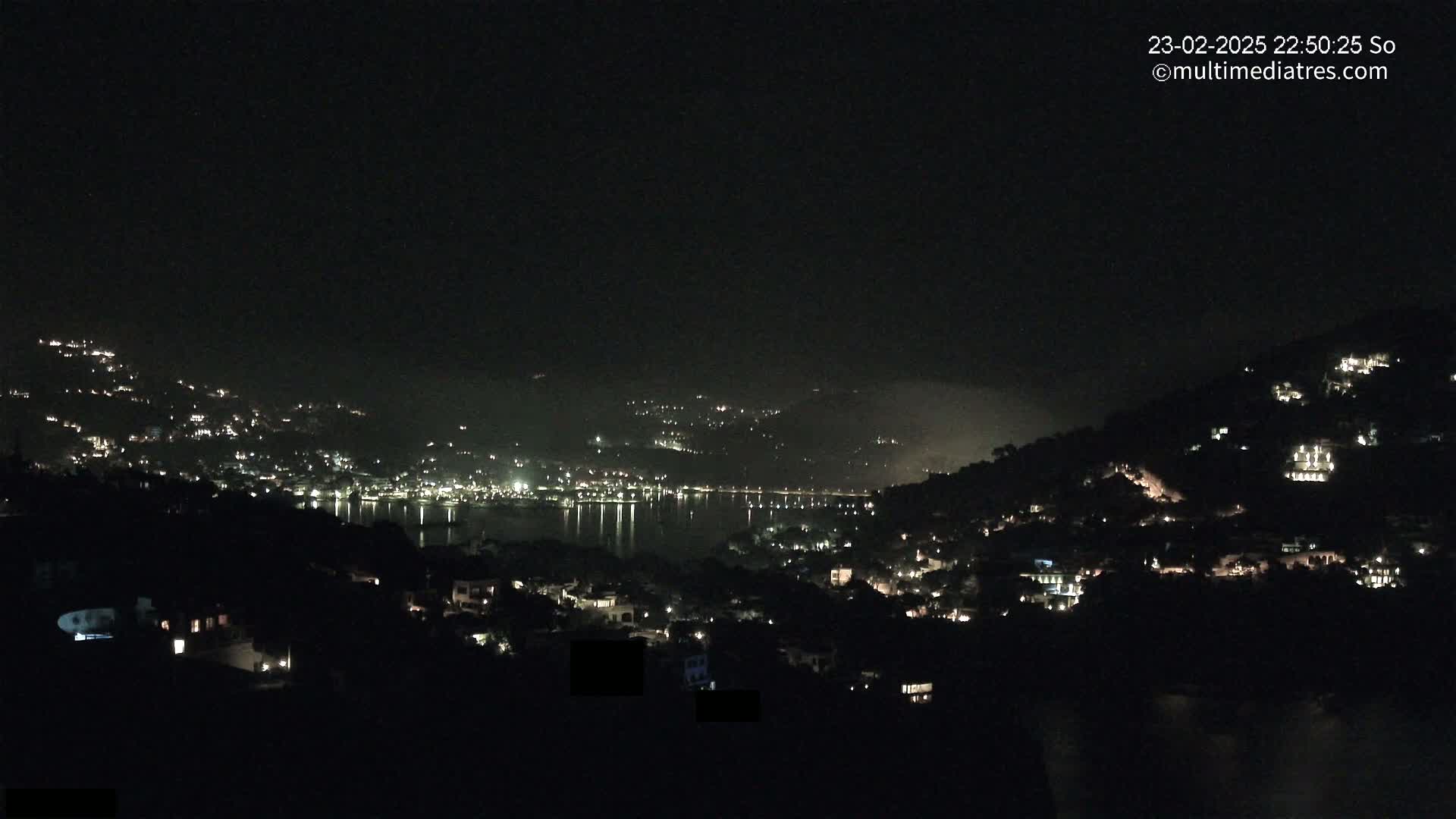 A nighttime view shows a body of water reflecting the lights of a town nestled on hillsides under a dark, clear sky.
