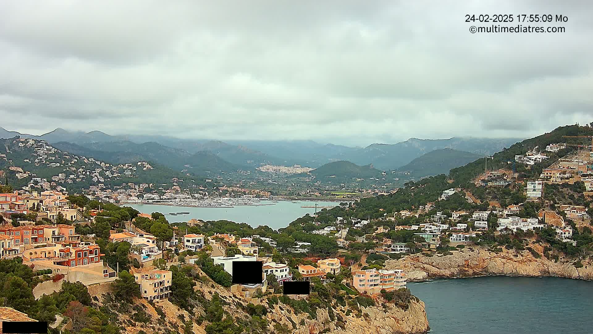 A coastal town nestled in a mountainous region under a cloudy sky overlooks a calm bay filled with boats.