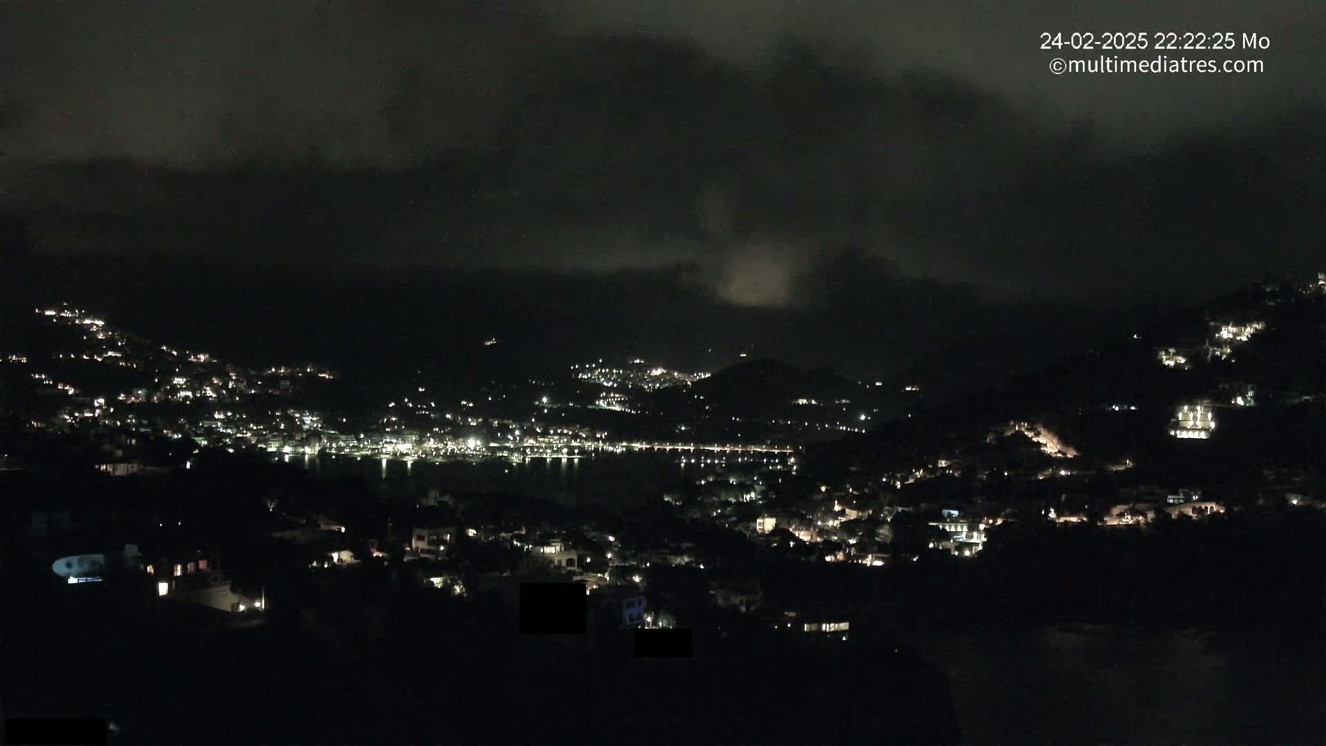 A nighttime view of a coastal town nestled on hillsides under a cloudy sky, with the town lights illuminating the landscape.
