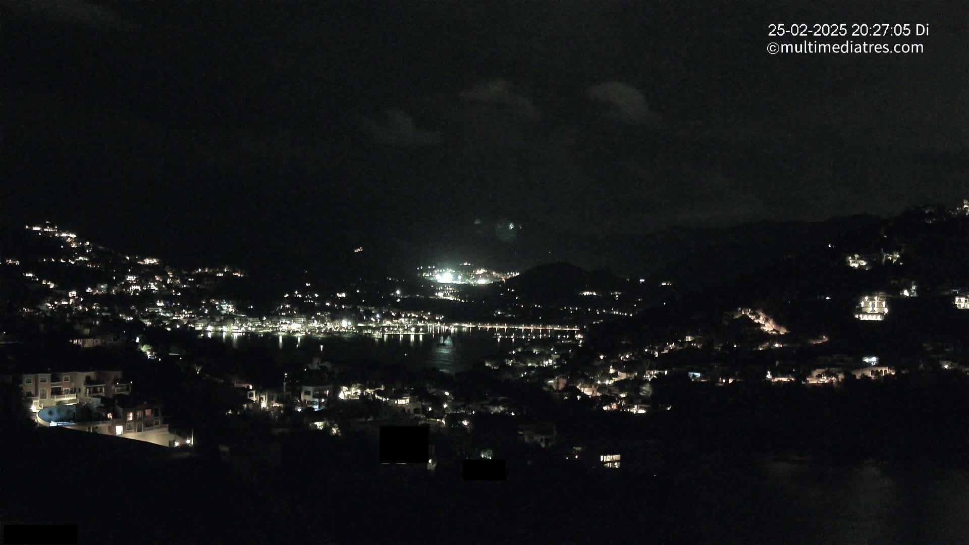 A nighttime view of a town nestled on a hillside overlooking a dark body of water under a partly cloudy sky.