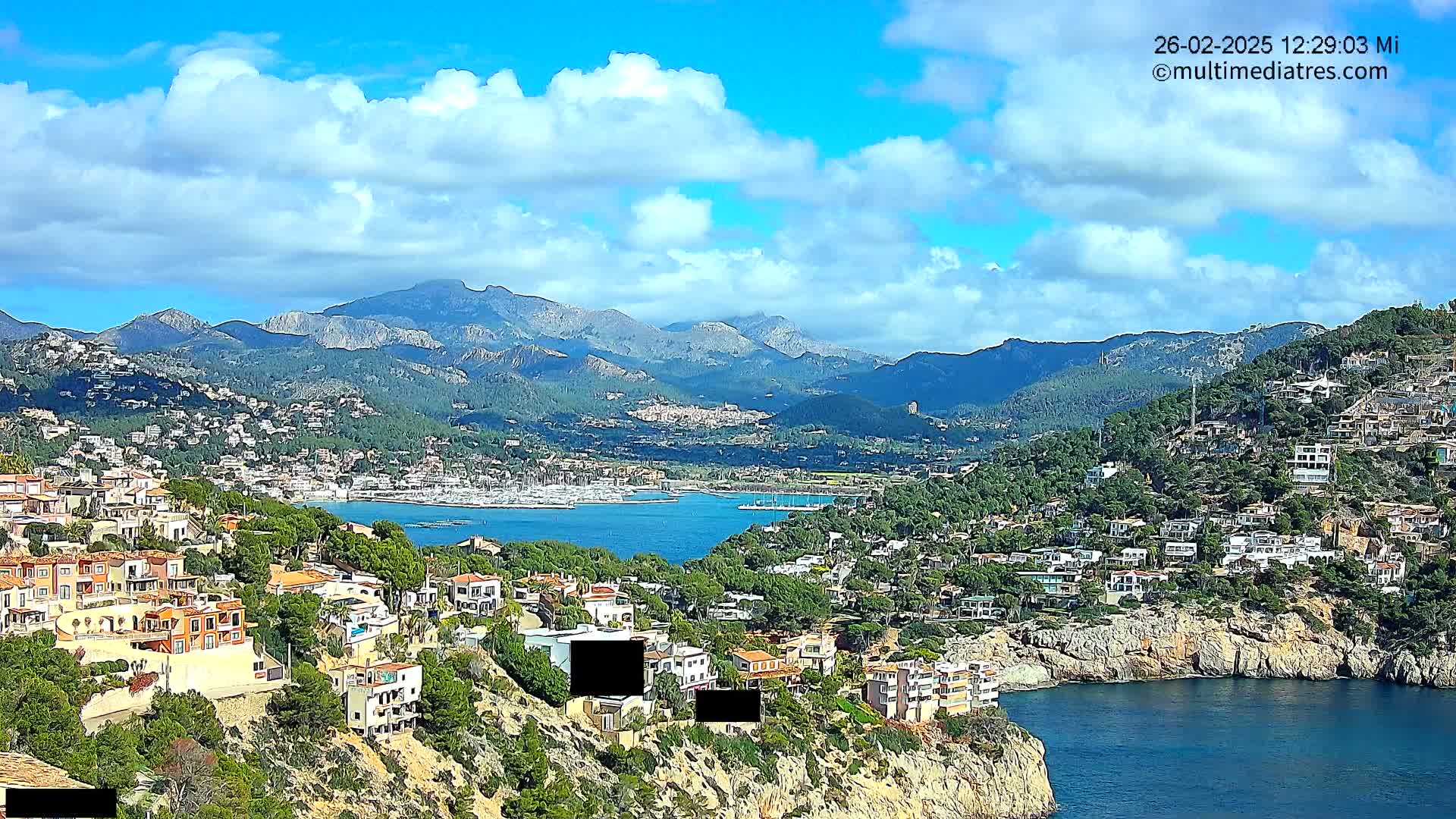 A coastal town nestled in a mountainous region with a harbor full of boats under a partly cloudy blue sky.