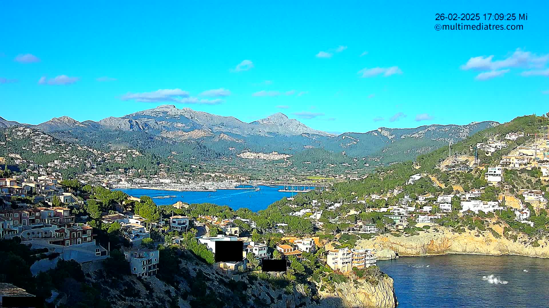 A coastal town nestled against a mountain range under a clear blue sky, with a calm body of water containing several boats in the foreground.