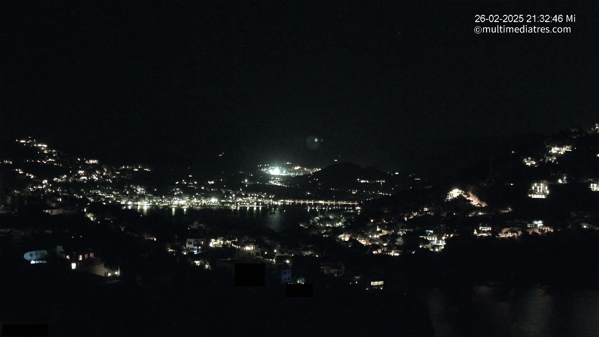 A nighttime view of a coastal town nestled on hillsides, with scattered lights illuminating houses and a body of water.