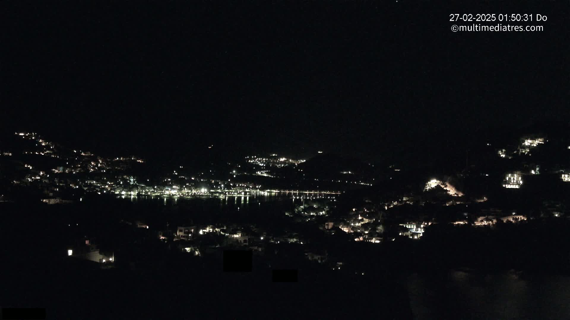 A nighttime view of a coastal town illuminated by scattered lights, nestled against dark hills and a calm body of water under a clear, dark sky.