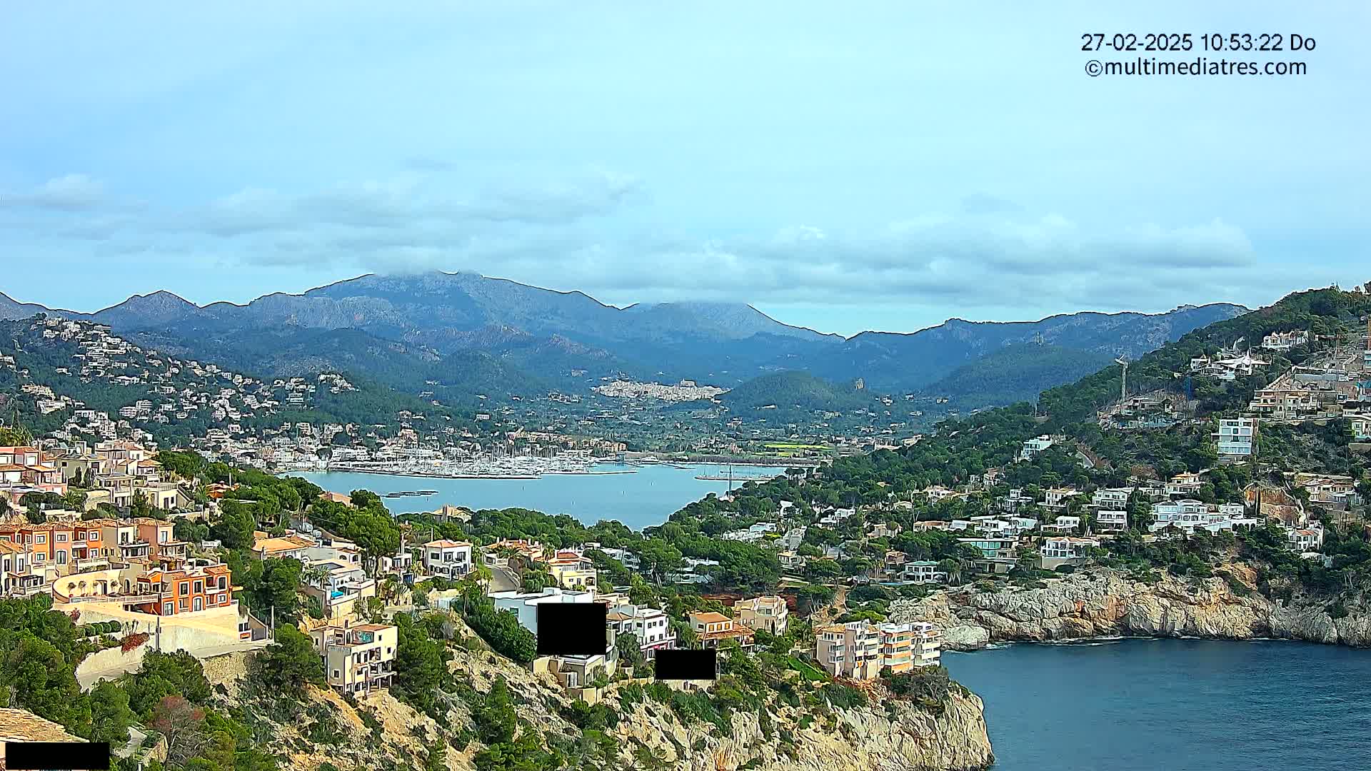 A coastal town nestled on a hillside overlooking a calm bay with a marina and mountains in the background under a partly cloudy sky.