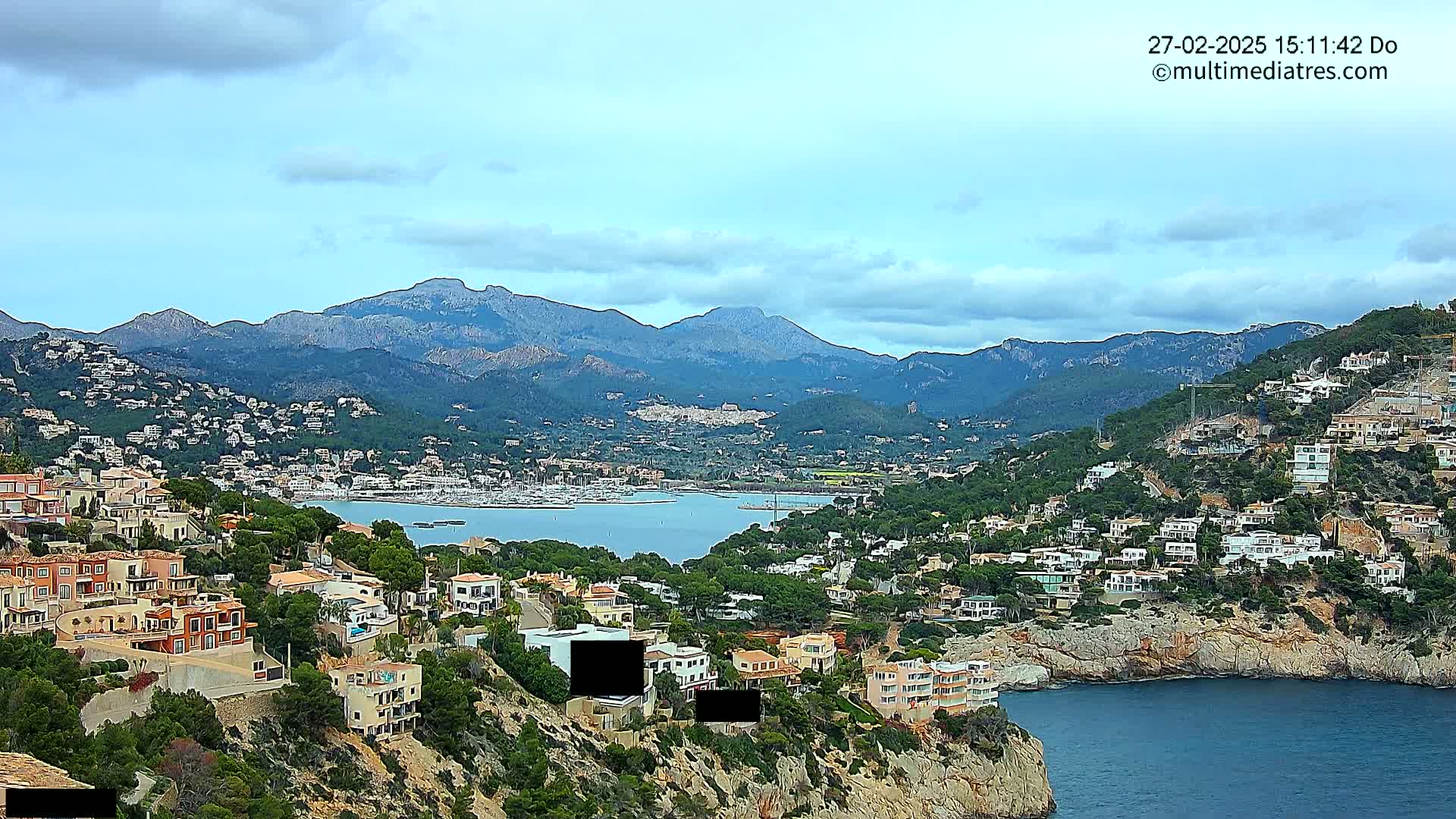 A coastal town nestled into a hillside, overlooking a calm bay with many boats and a mountainous backdrop under a mostly cloudy sky.