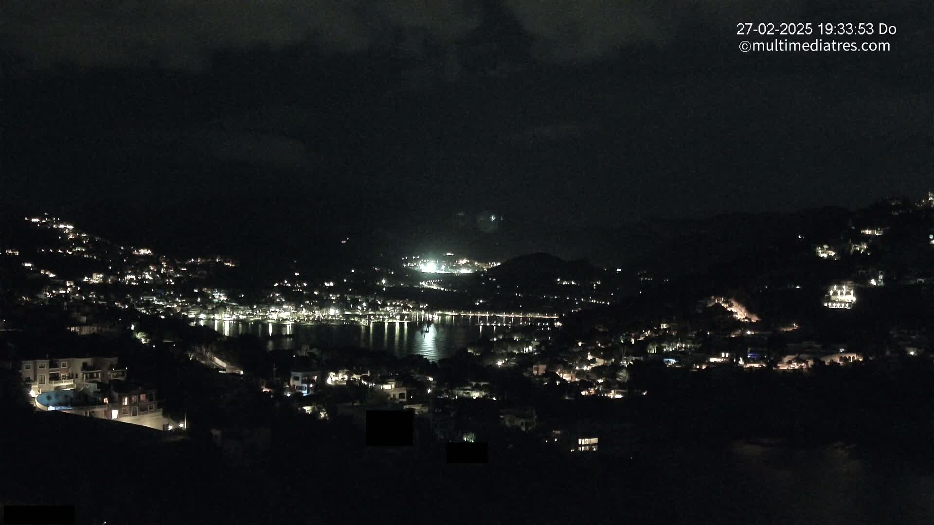 A nighttime view of a coastal town illuminated by many lights, with a calm body of water reflecting the lights under a partly cloudy sky.