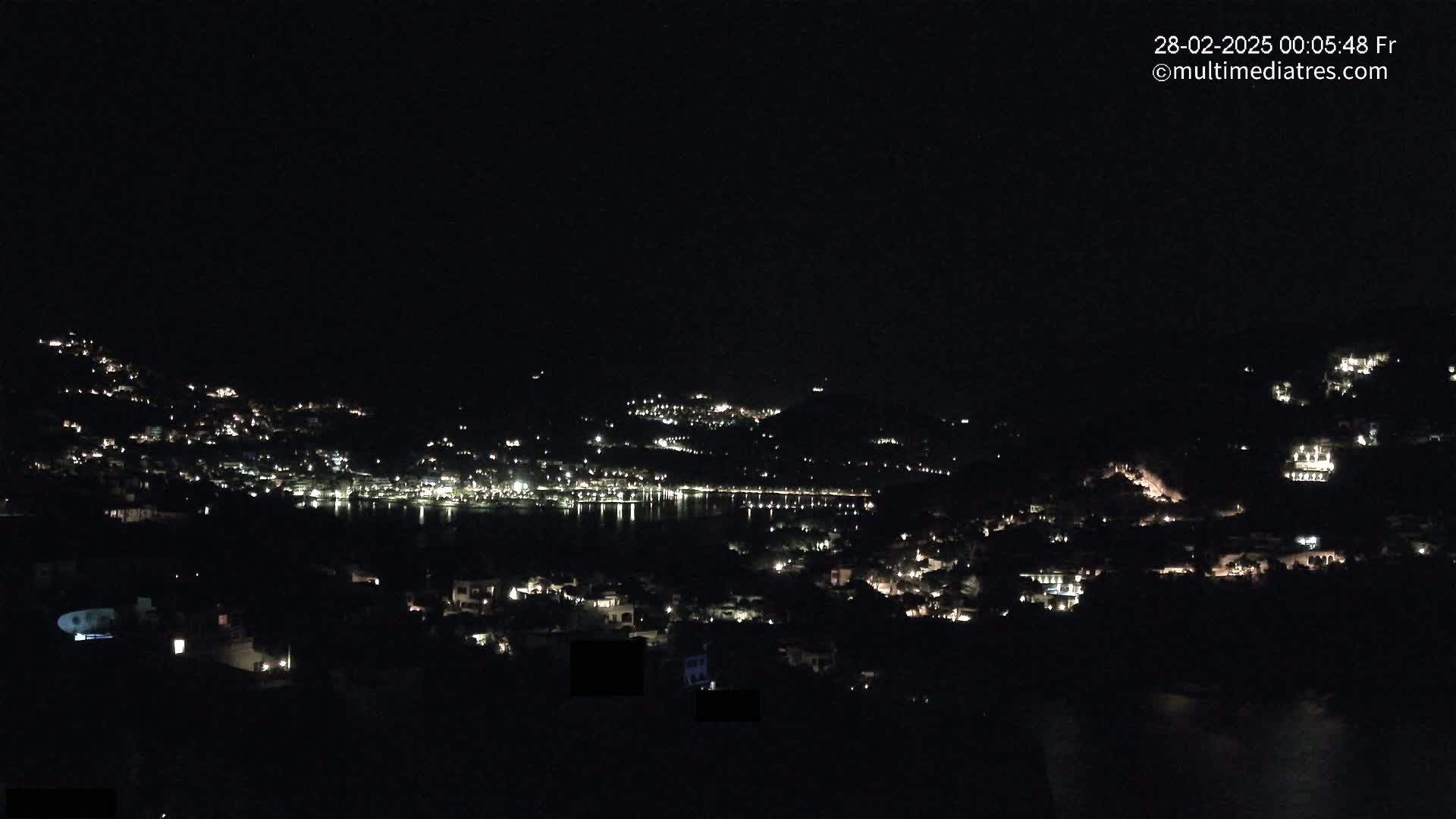 A nighttime view of a coastal town nestled on hillsides, with scattered lights illuminating the buildings and reflecting on the dark water below.