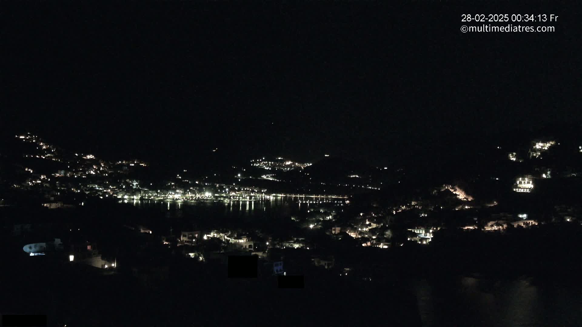 A nighttime view of a town nestled on hillsides surrounding a dark body of water, with scattered lights illuminating the buildings and reflecting faintly on the water.