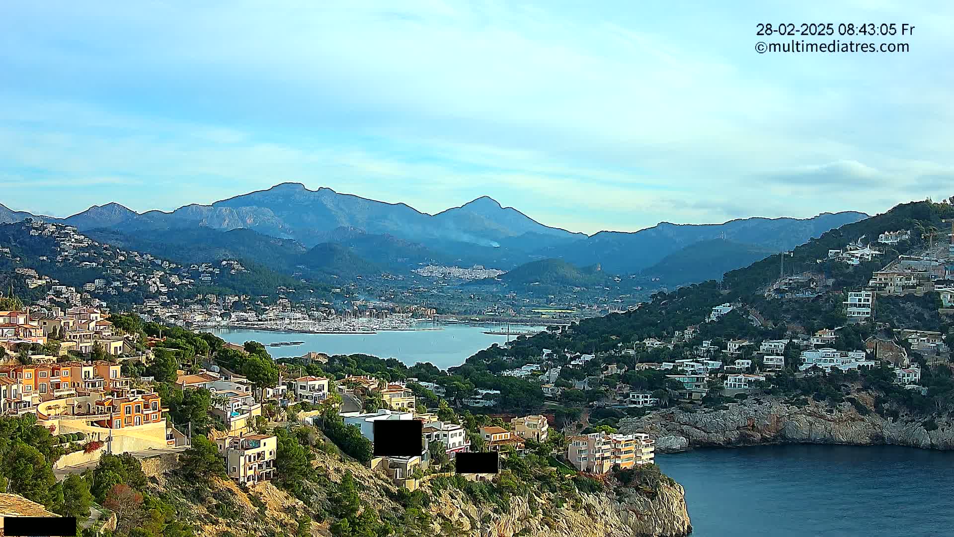A coastal town nestled on a hillside overlooking a calm bay filled with boats, with mountains in the background under a clear, bright sky.