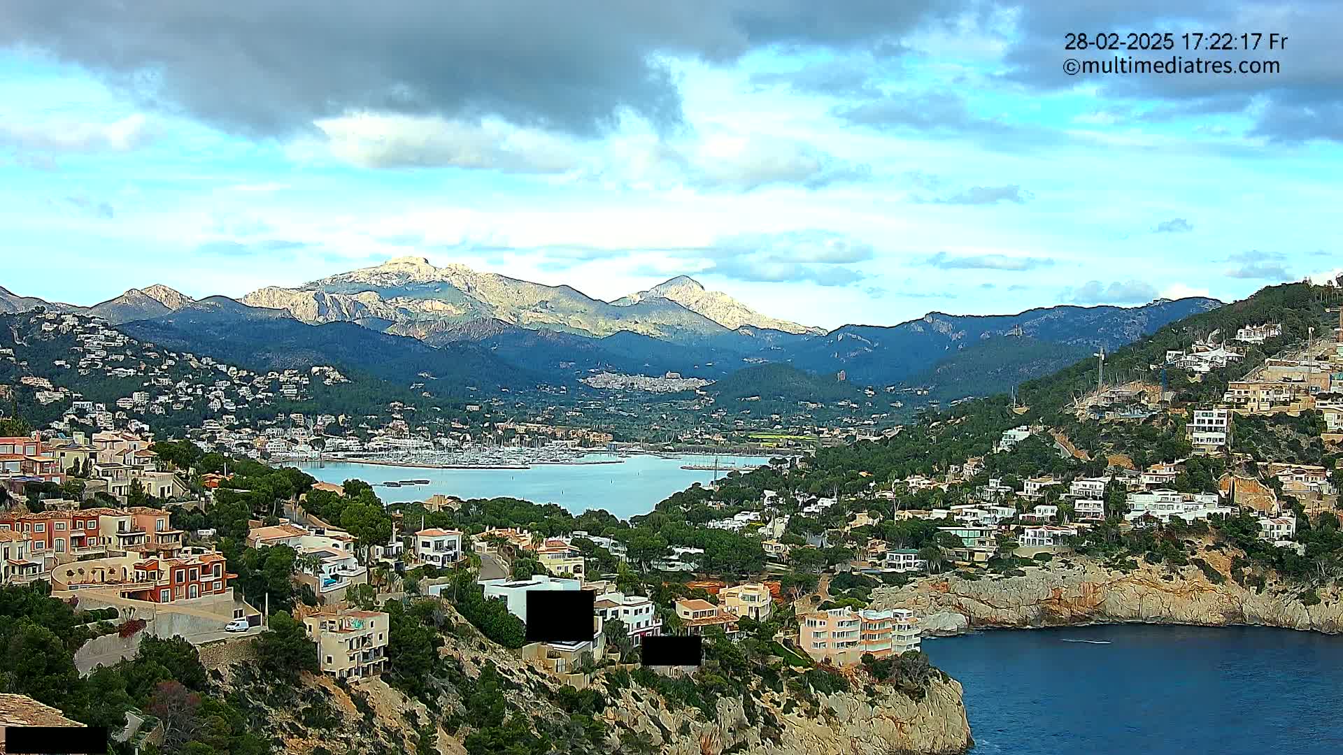 A coastal town nestled between a calm bay and mountains under a partly cloudy sky.