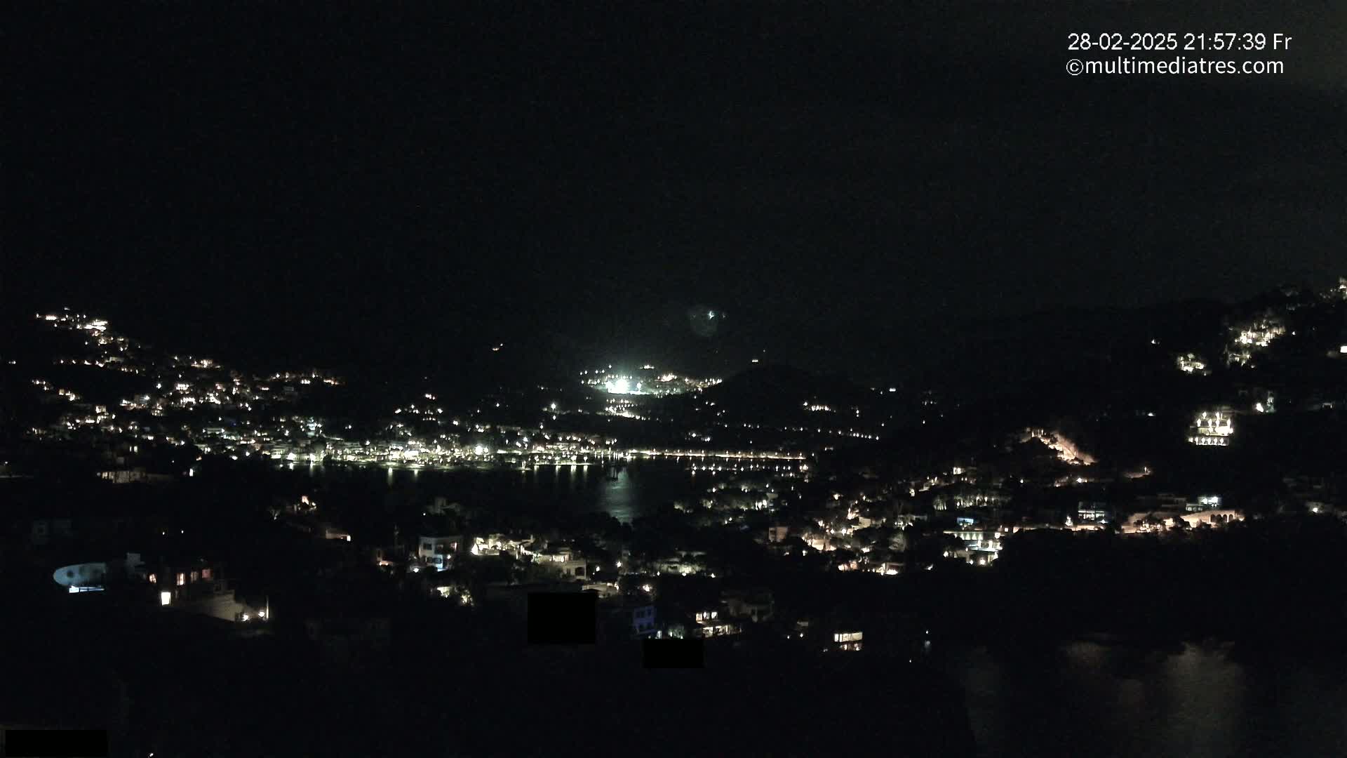 A nighttime view of a coastal town illuminated by scattered lights, situated on a dark hillside overlooking a body of water under a clear, dark sky.