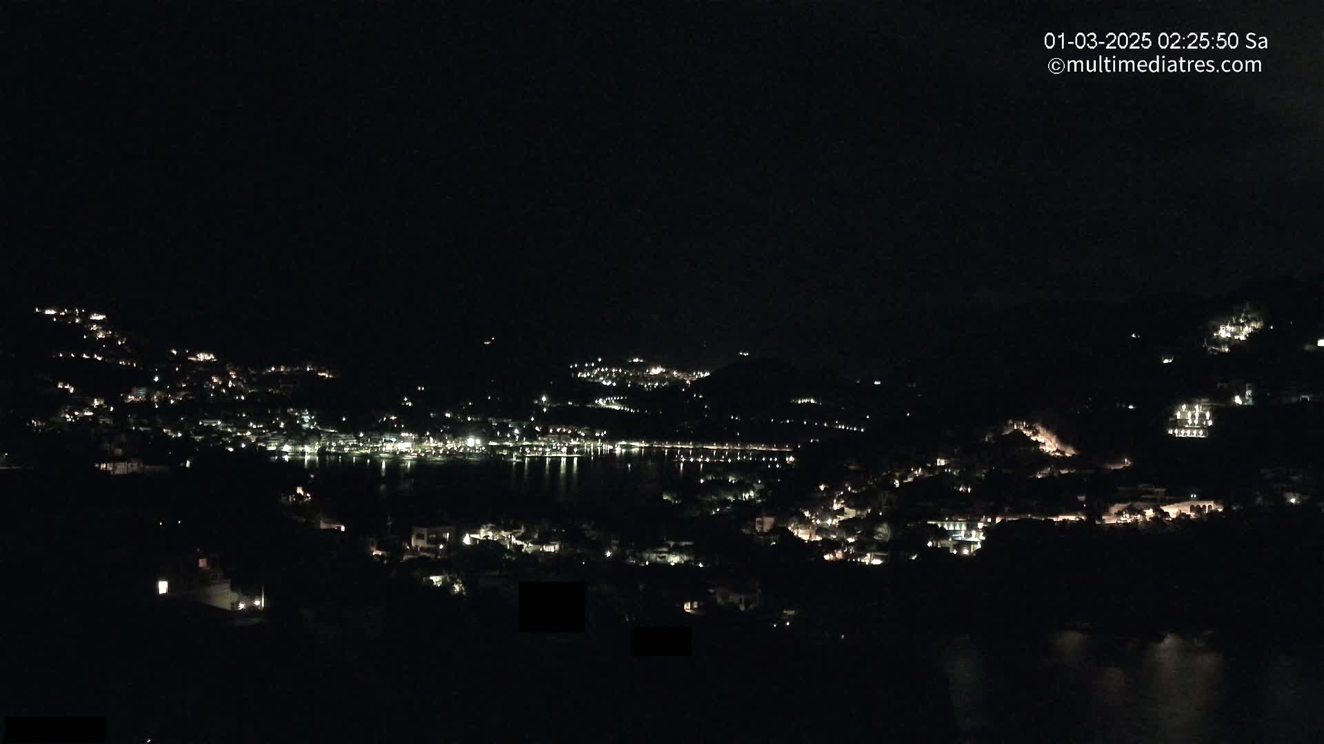 A nighttime view of a town nestled on hillsides surrounding a dark body of water.