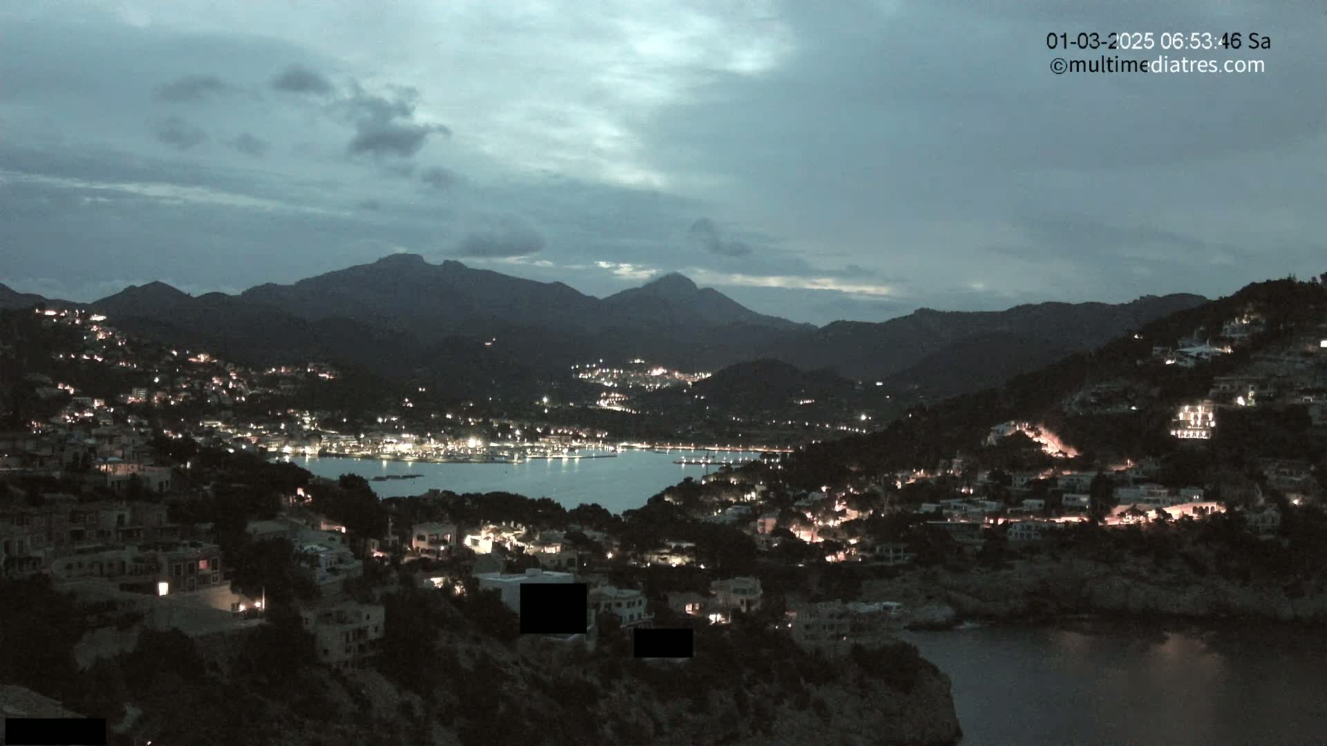 A nighttime view of a coastal town nestled between mountains, illuminated by scattered lights under a cloudy sky.
